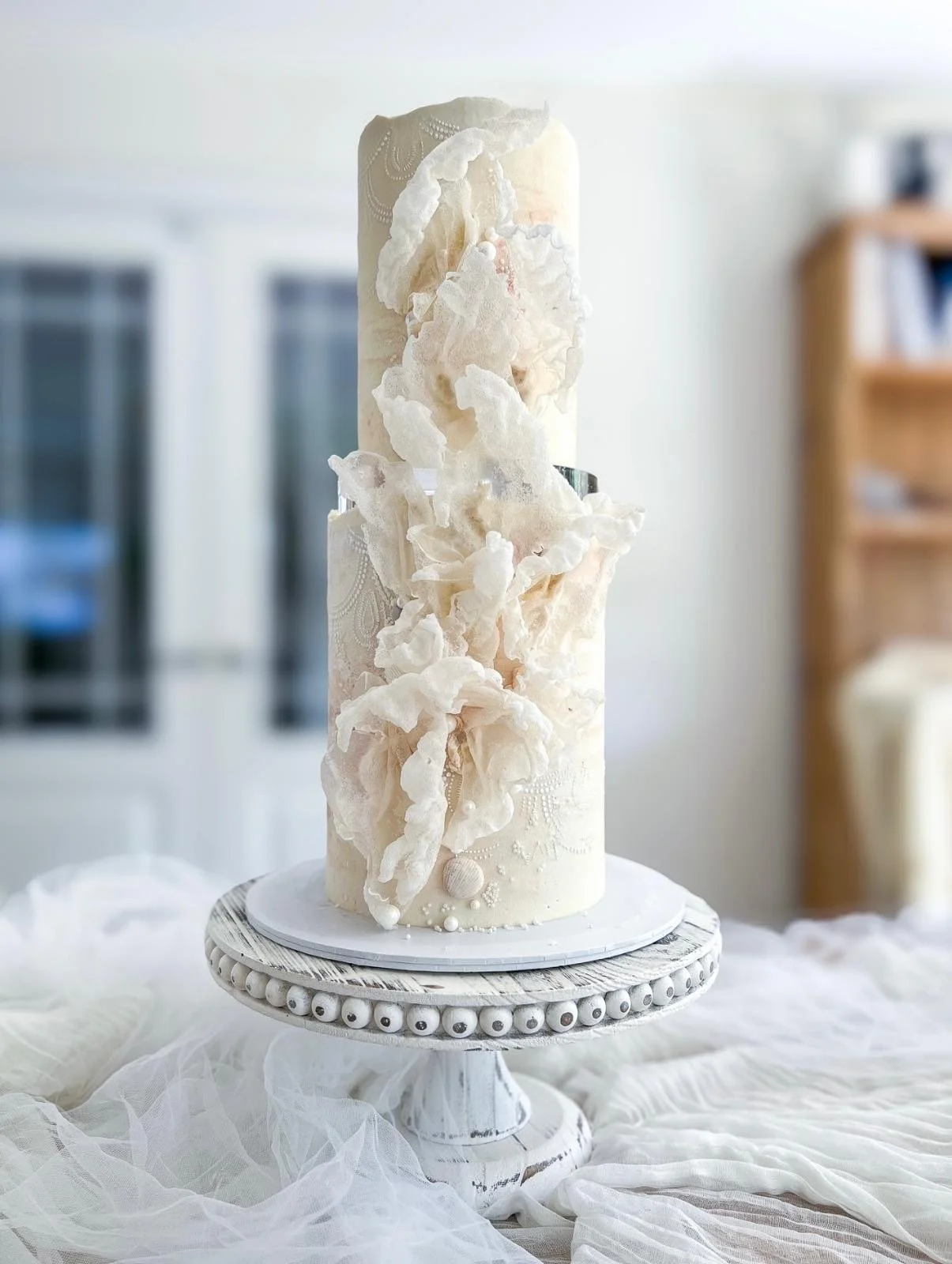 A tall white wedding or special occasion cake with intricate textured fondant and decorative elements, displayed on a white pedestal cake stand in a bright room.