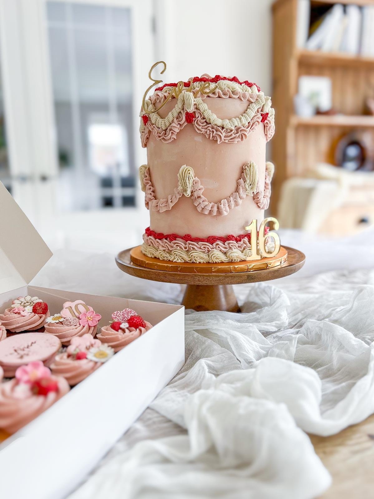 Pink birthday cake with white and red frosting ruffles, a gold "Love" topper, and a gold "16" at the base, on a wooden cake stand. A box of pink and red decorated cupcakes with flowers and berries is in the foreground. The background includes a window and a bookshelf.