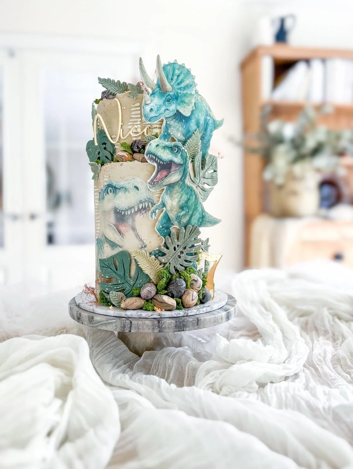 Decorative cake with dinosaur-themed toppers, greenery, and stones, displayed on a wooden stand with white fabric surrounding it, in a bright indoor setting.