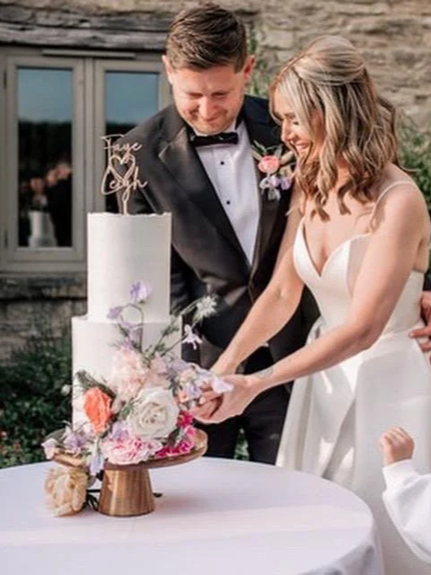 A bride and groom cutting a wedding cake together outdoors, with the bride in a white dress and the groom in a black tuxedo, surrounded by flowers and a rustic building in the background.