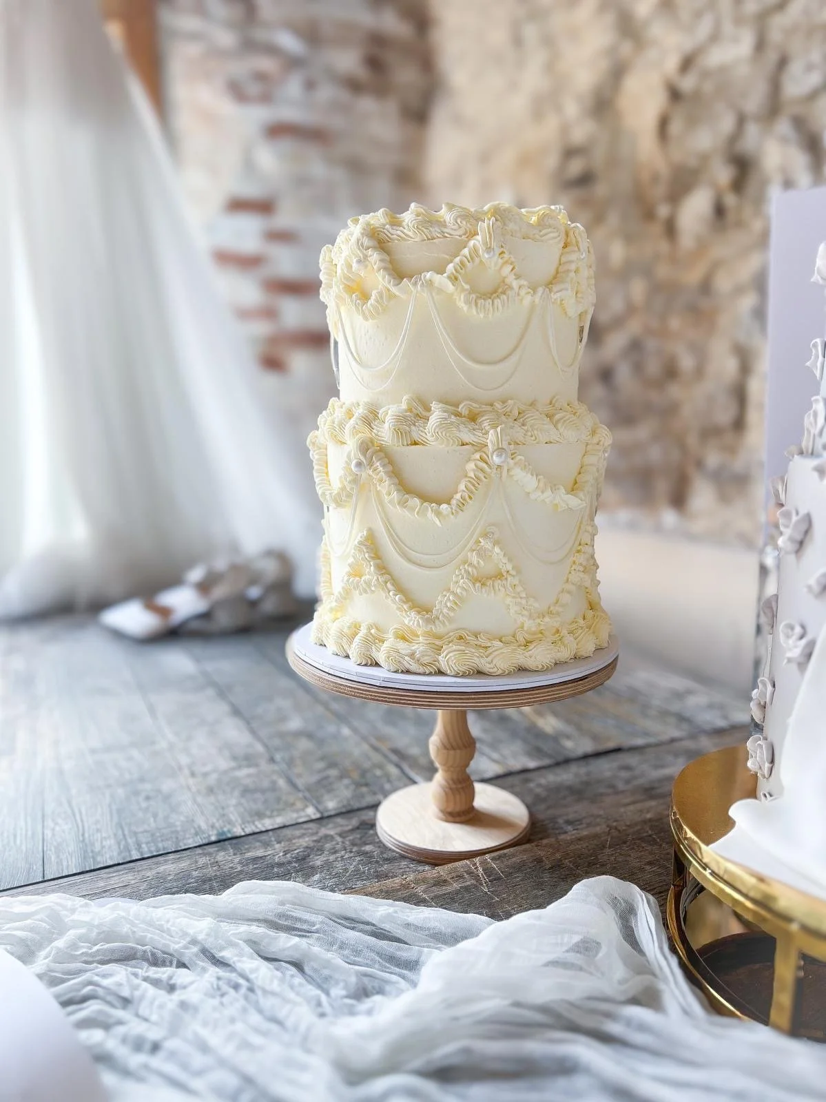 A decorated wedding cake with two layers, cream-colored frosting, piped swags, pearls, and ruffles, placed on a wooden cake stand on a rustic wooden table.