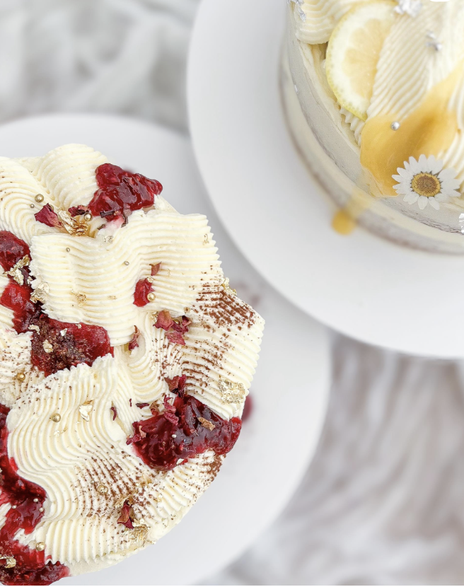 Close-up of two decorated cakes on white plates with a wooden table background. One cake is topped with white buttercream, raspberry sauce, edible gold flakes, and gold sprinkles. The other cake has lemon slices, white frosting, and daisy edible decorations.