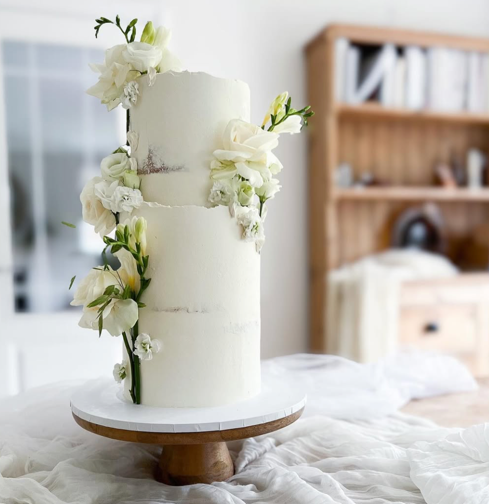 A tall, double-tiered white wedding cake decorated with white flowers, placed on a wooden cake stand in a cozy indoor setting.