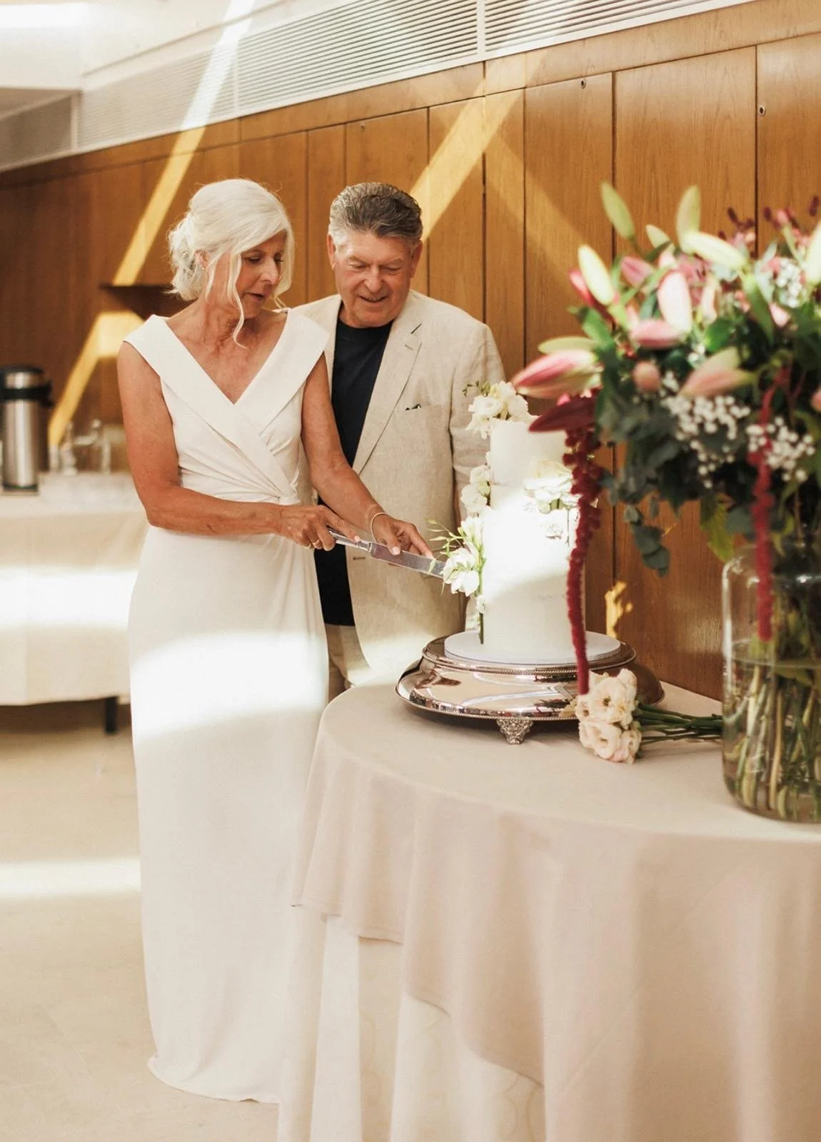 A middle aged couple at a wedding celebration, with the woman in a white dress and the man in a light jacket, cutting a white wedding cake together, with a large bouquet of pink and white flowers nearby.