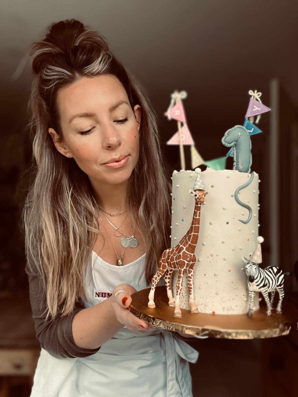 A woman with long wavy hair, wearing a white apron and necklaces, holding a cake decorated with animal figures including a giraffe, zebra, and elephant, with colorful bunting on top.
