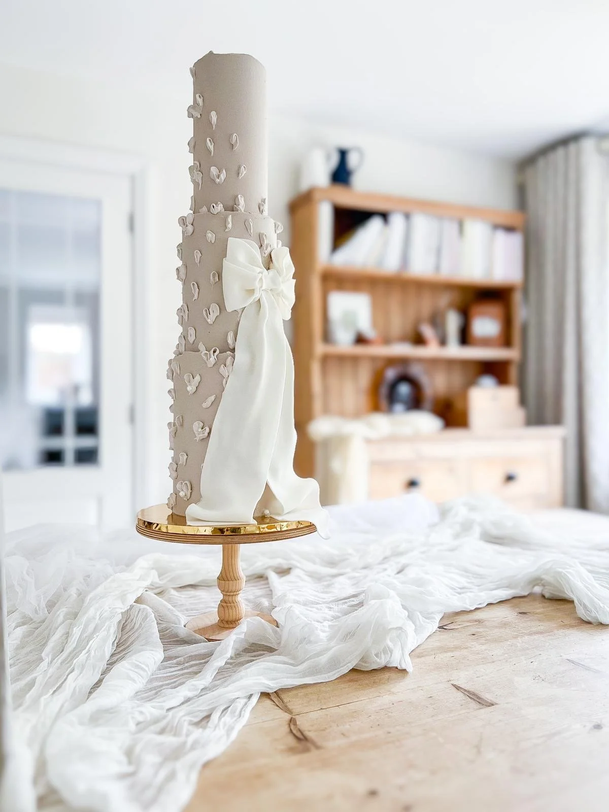 A tall, cylindrical wedding cake decorated with small white flower appliques and a large white bow, placed on a round wooden stand on a table covered with white fabric, in a cozy room with wooden shelves and a door in the background.