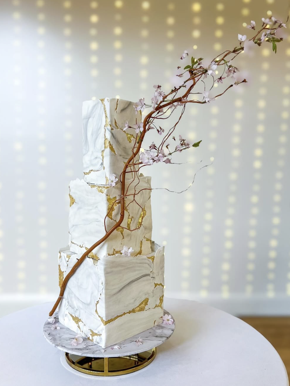 Elegant multi-tiered cake with white and gold marbled design, topped with a branch of pink blossoms, set on a white table with a decorative base.