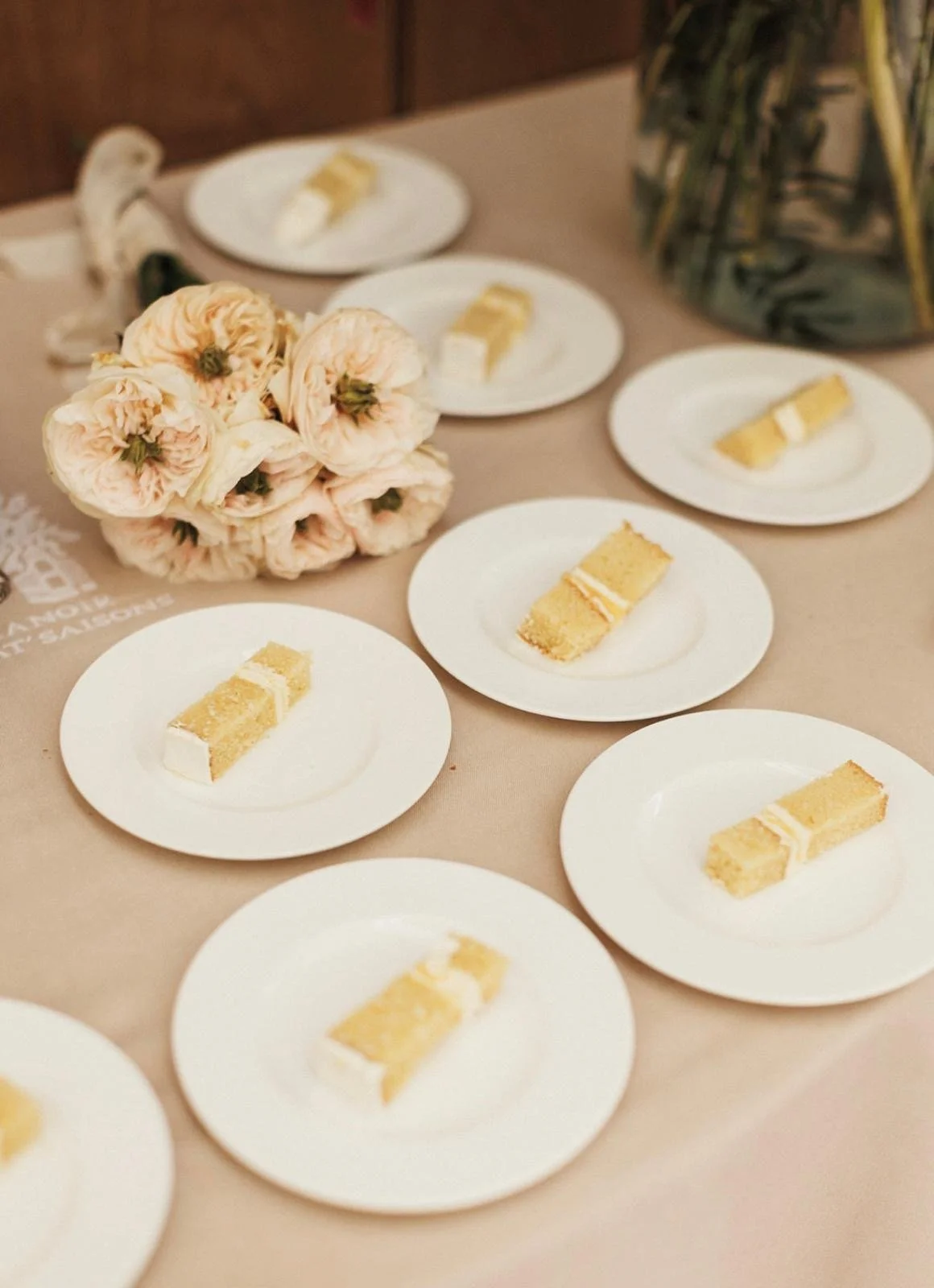 A table with multiple white plates, each holding a piece of layered yellow and white cake. In the center, there is a bouquet of light pink roses and a large glass jar filled with green leafy plants.