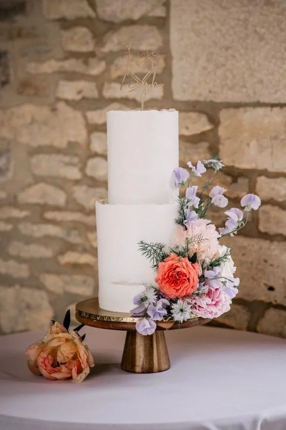 Two-tiered white wedding cake adorned with pink and purple flowers on a wooden cake stand, with a peach-colored flower lying on the table nearby, against a stone wall background.