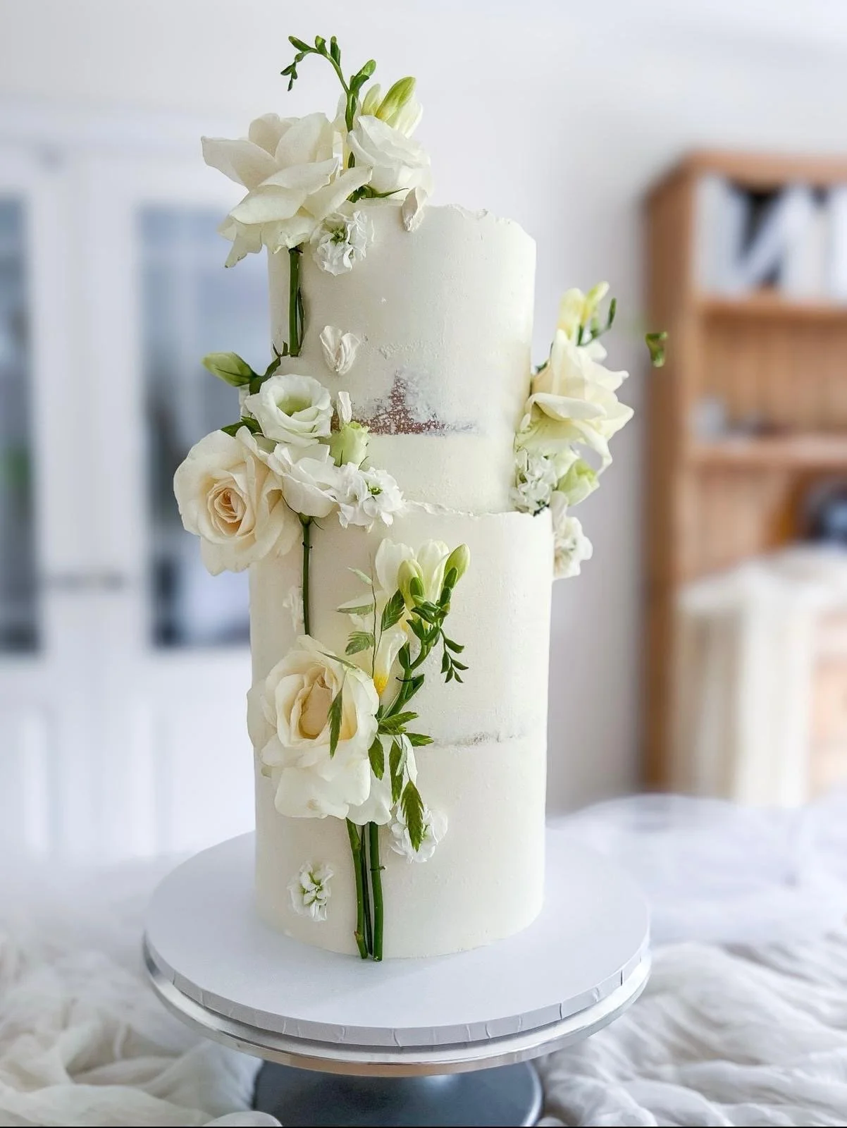 A three-tiered white wedding cake decorated with white flowers and green leaves, displayed on a white cake stand.