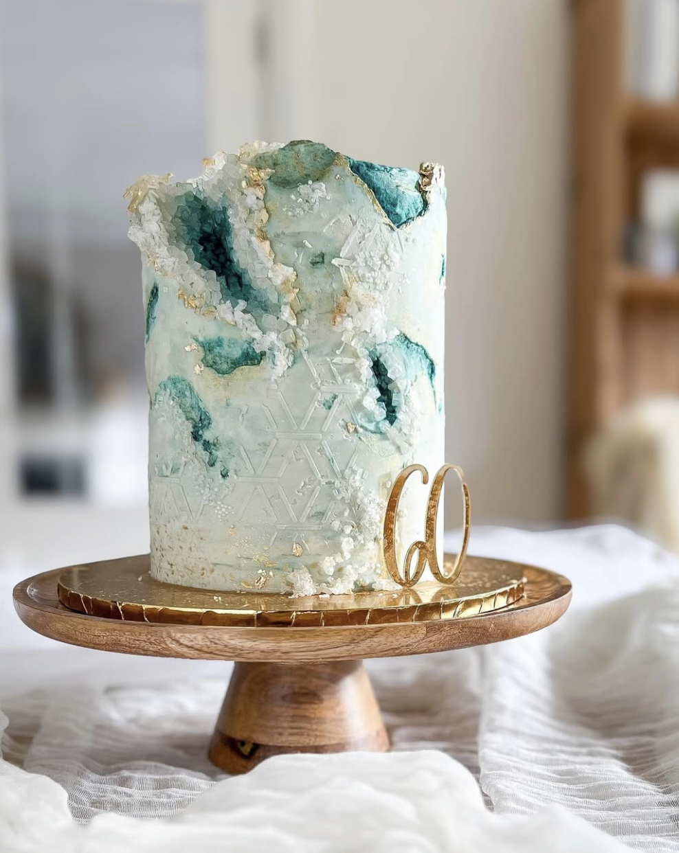 A tall, artistic birthday cake with an icy, glacier-like appearance, decorated with gold accents and a gold letter 'd' cake topper, displayed on a wooden cake stand.