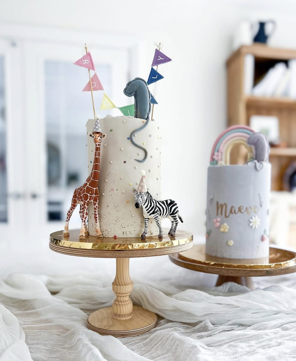 Two birthday cakes on wooden stands with animal and rainbow decorations. The larger cake has giraffe and zebra figures, small colorful flags, and a number one topper. The smaller cake is decorated with a rainbow and the name 'Maeve' and has pastel co