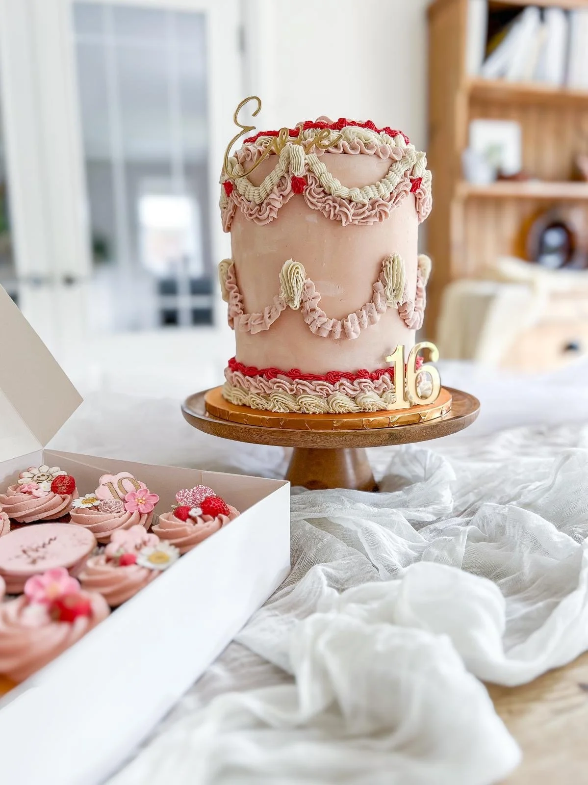 A two-tiered pink birthday cake with cream-colored and red frosting decorations, a gold "love" cake topper, and the number 16 is on the cake stand. There is a white box of pink decorated cupcakes with raspberries and flowers in the foreground.
