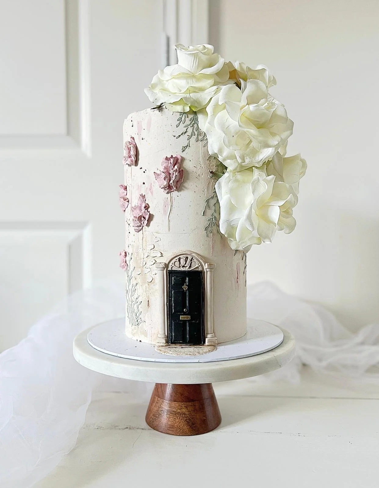 A tall, elegant cake decorated with white and pink flowers, featuring a black door ornament at the bottom, placed on a white stand.