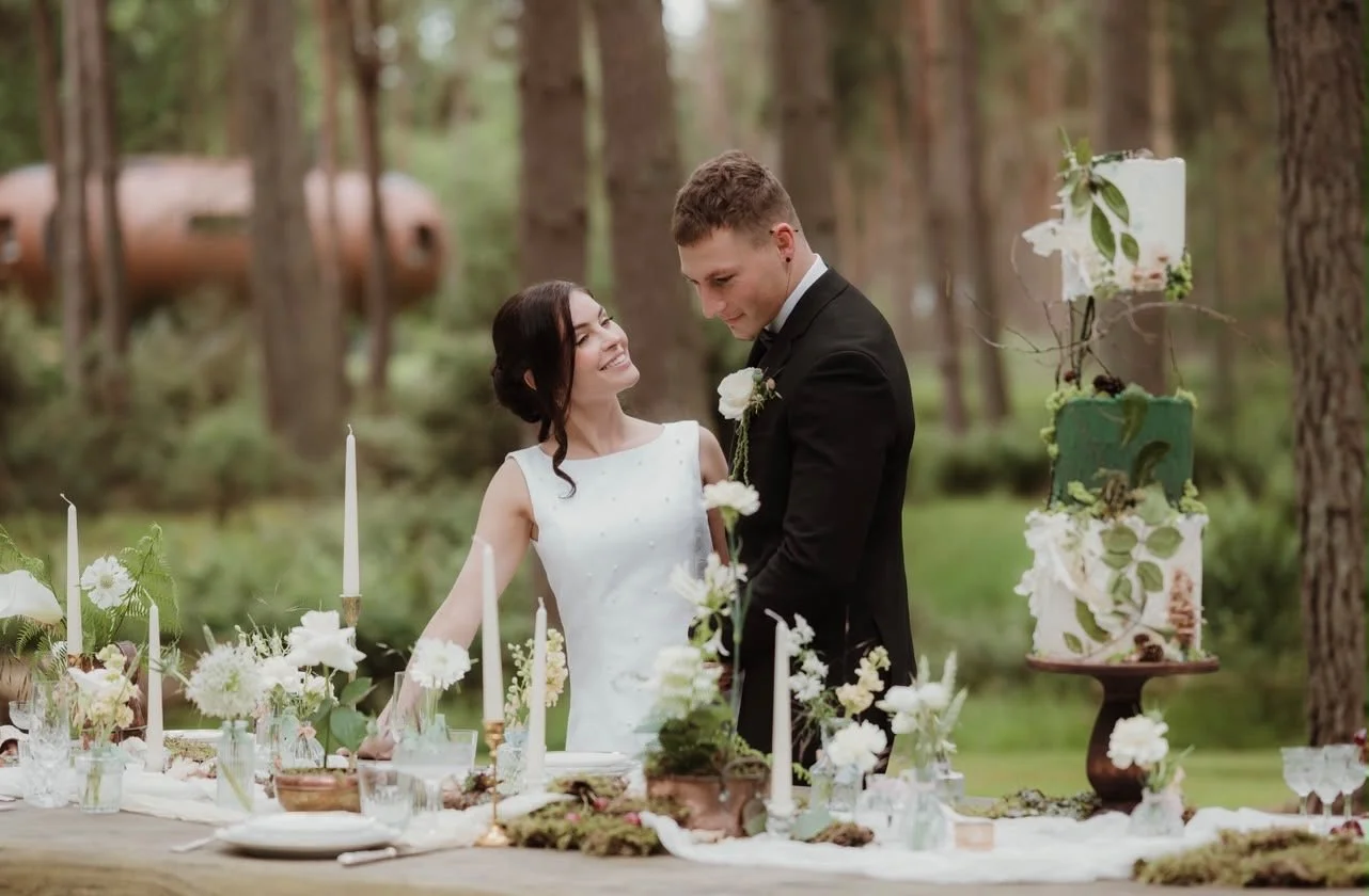 A bride and groom standing together at an outdoor wedding reception, smiling at each other, with decorated tables and a multi-tiered cake in a wooded area.