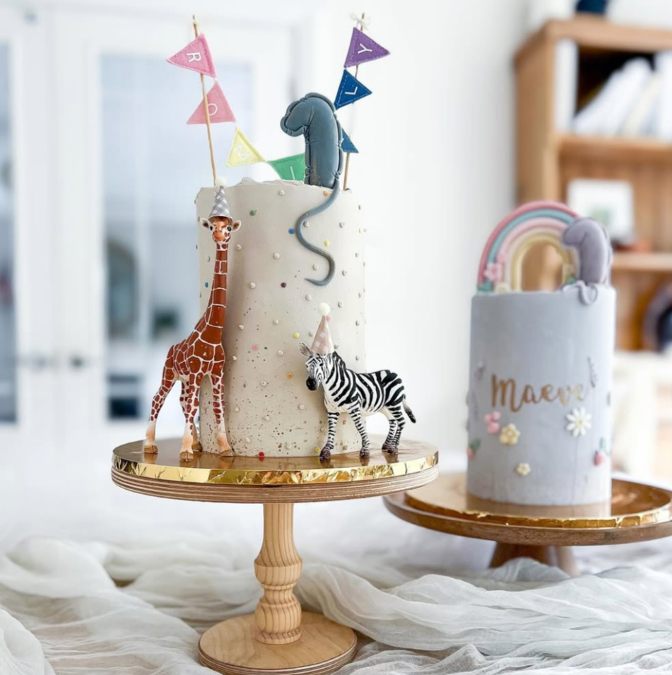 Two decorated birthday cakes on wooden stands, featuring animal and rainbow themes, with colorful bunting and garnishes.