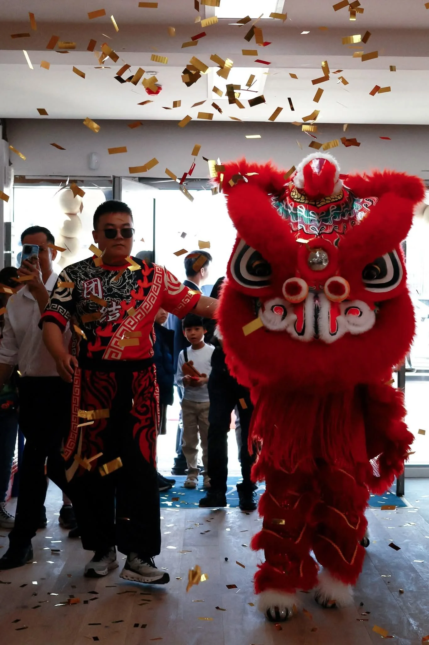 A lion dance performer in a red costume with a traditional lion head mask, standing in front of a group of people during a celebration with falling gold and red confetti.