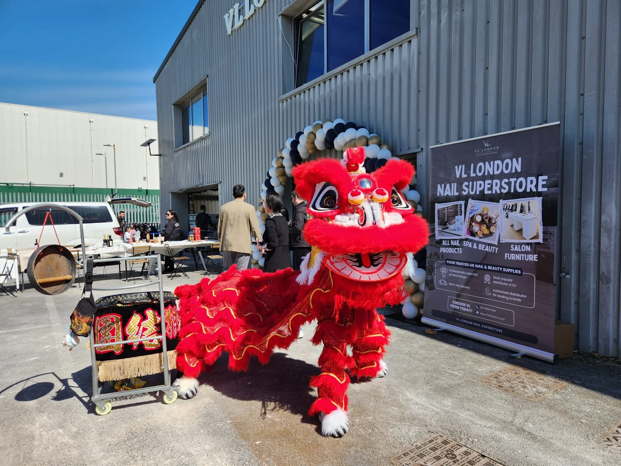 A bright red lion dance costume with white and black accents standing outdoors in front of a balloon arch and a sign for VL London Nail Superstore. People are gathered in the background near tables and a building with metal siding.