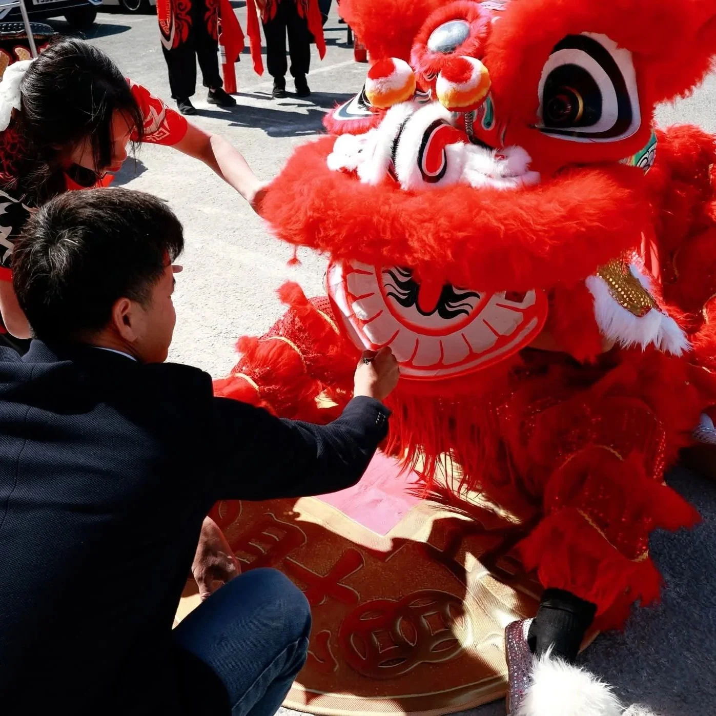 People gather around a red Chinese lion costume, preparing for a lion dance performance. Two individuals are adjusting or painting the lion's face, while others are in the background. The setting appears to be an outdoor area with cars visible.