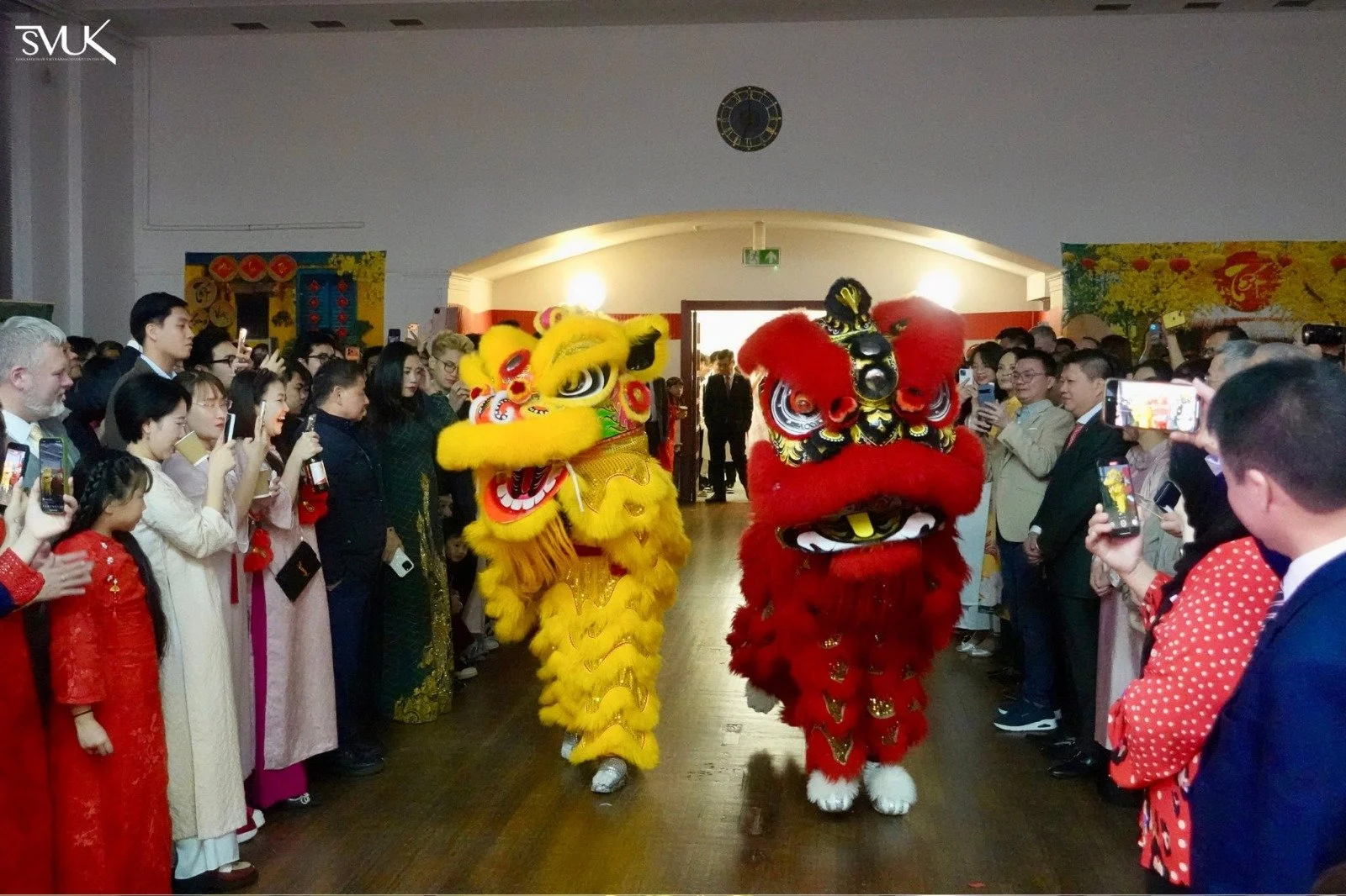 Lion dance performers in yellow and red costumes perform for a large audience at an indoor celebration, with many attendees dressed in traditional Asian attire, capturing the performance on their smartphones.