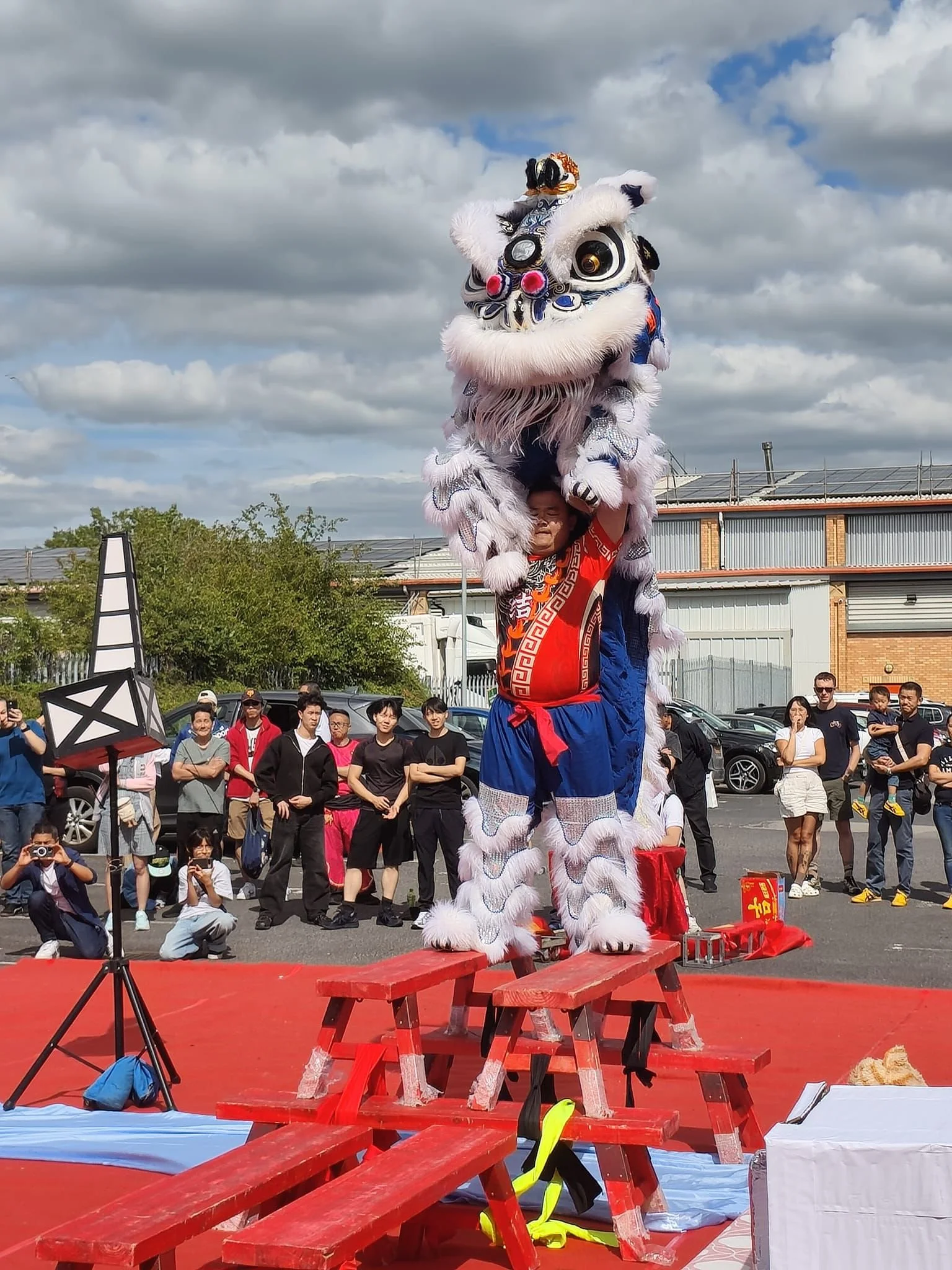 A performer in a traditional lion dance costume stands on a red platform during a festival or celebration. The costume is white with blue and black accents, large expressive eyes, and decorative elements. The performer raises the lion's head above their head while an audience watches.