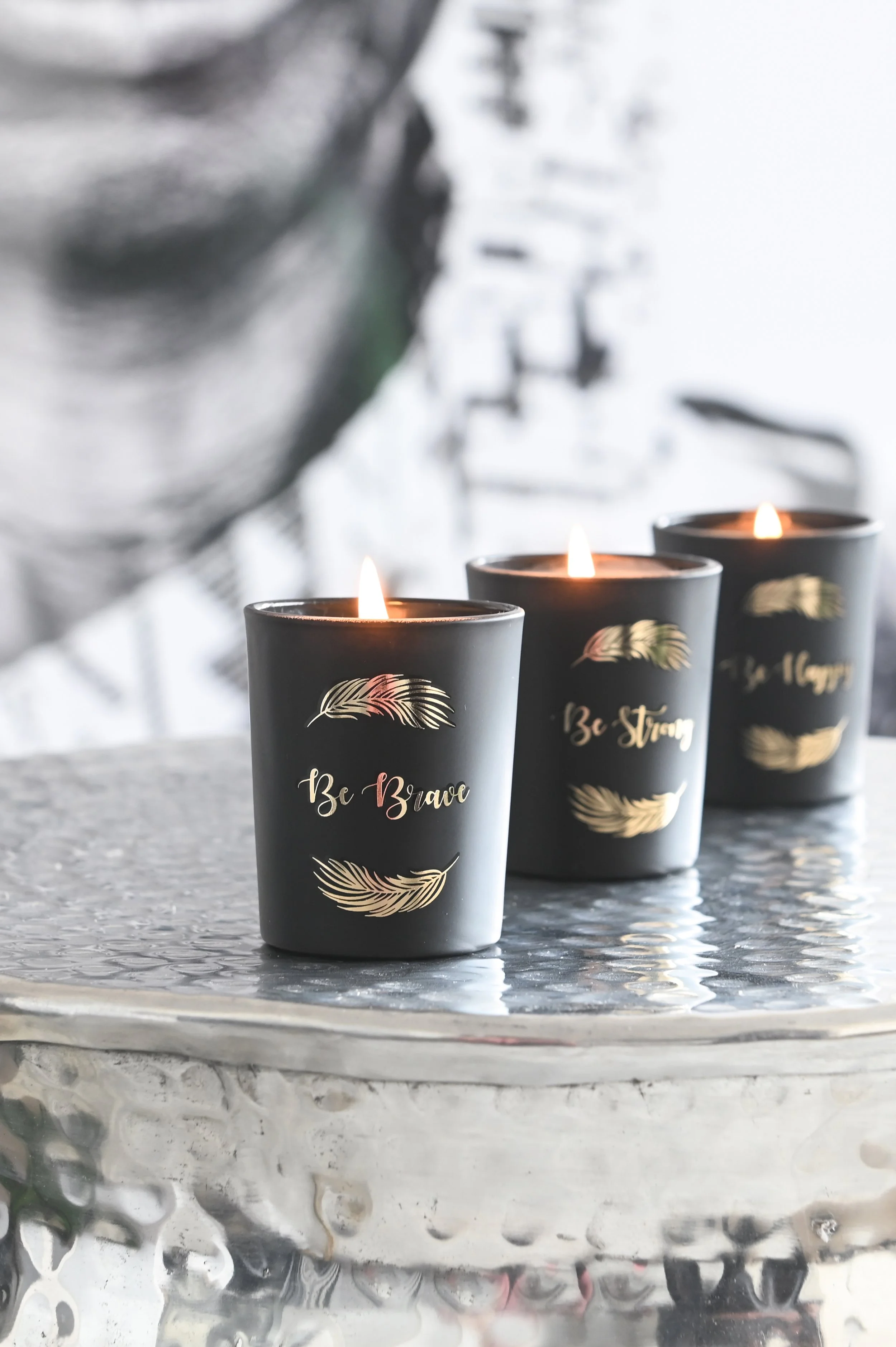Three black candles with golden feather designs and the words "Be Brave" and "Be Strong" written on them, lit and placed on a reflective silver surface, with a blurred background.