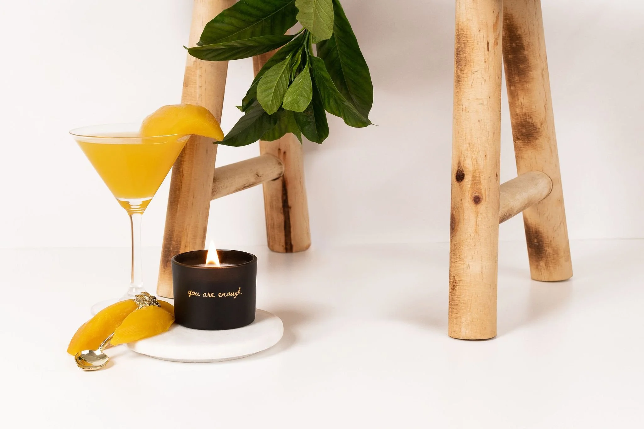 A black candle with gold writing that says 'you are enough', a small silver spoon, mango wedges and a mango daiquiri