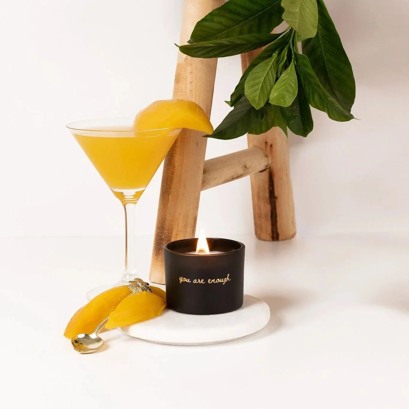 A black candle with gold writing that says 'you are enough', a small silver spoon, mango wedges and a mango daiquiri