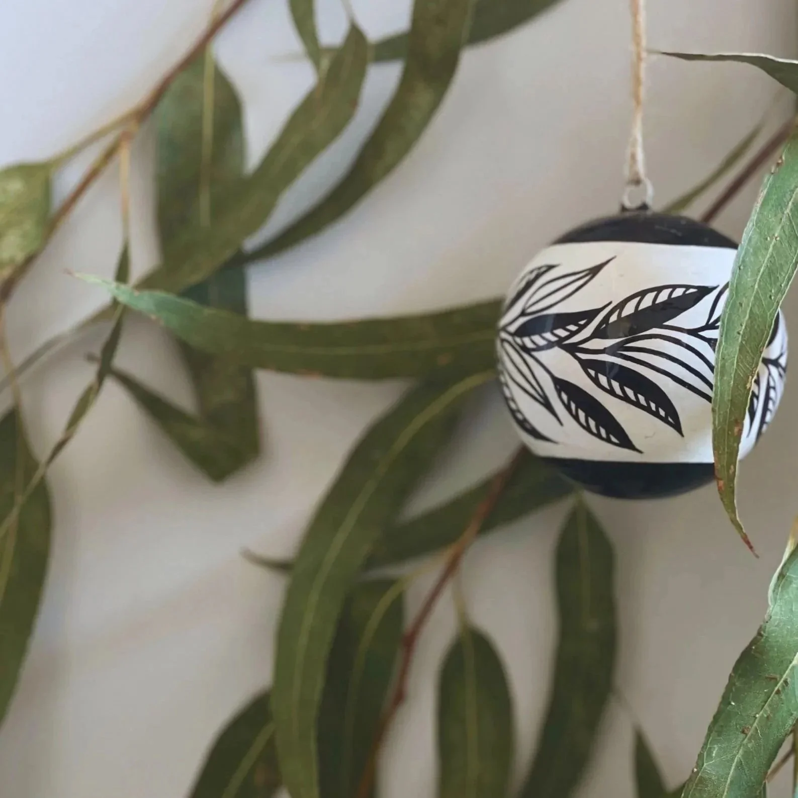 Decorative black and white ornament with a leaf pattern hanging amidst green eucalyptus leaves.