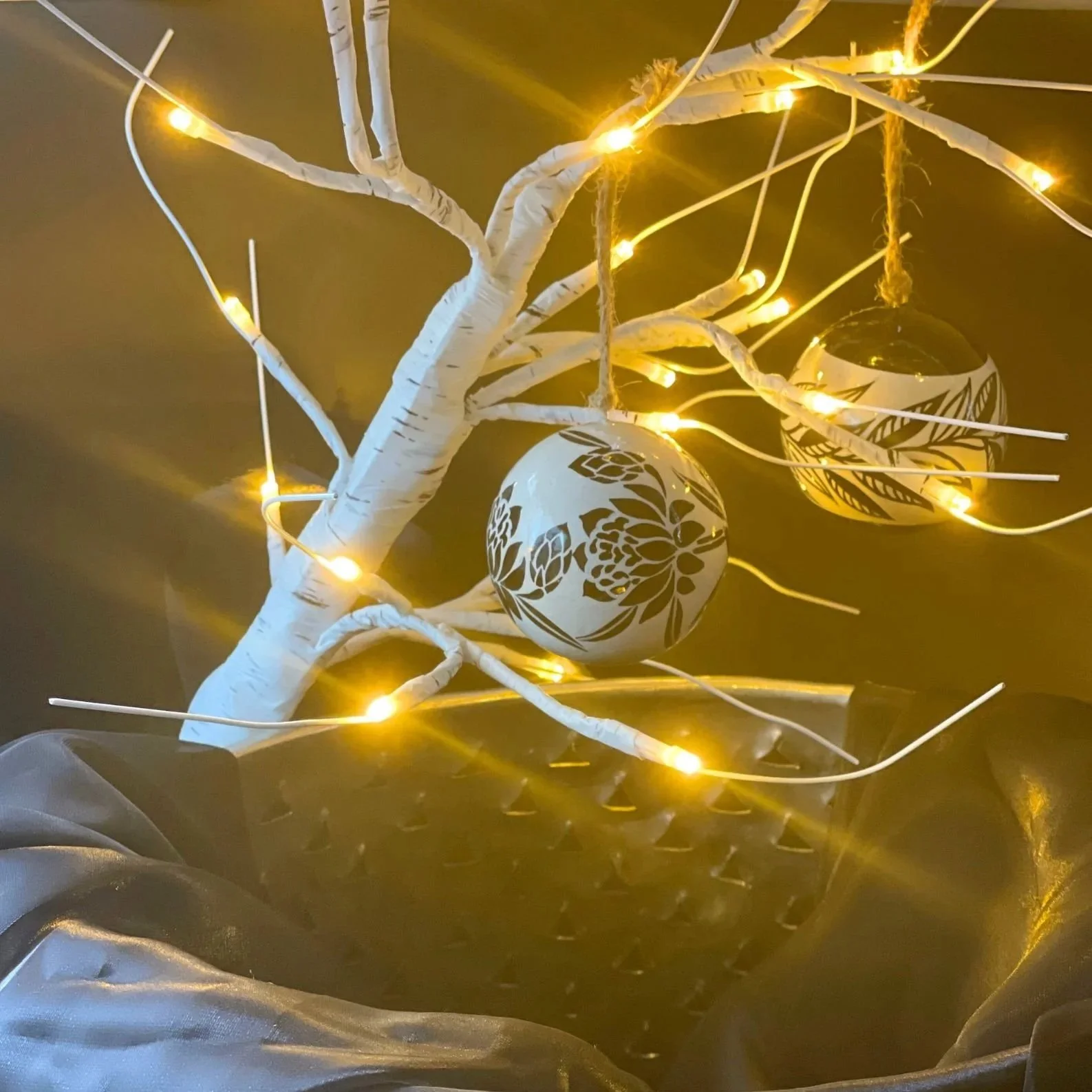 Decorative white tree branch with lights and hanging ornaments glowing in warm yellow light, set against a dark background.