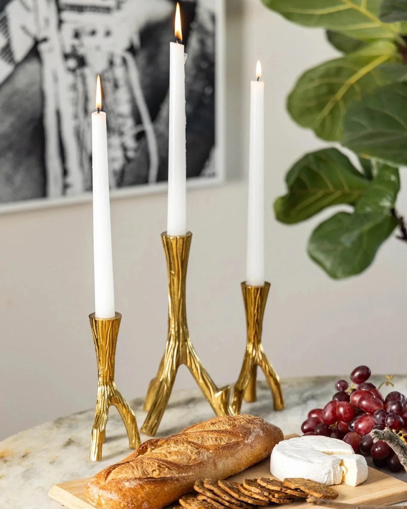 Gold candle stands with three lit candles, placed on a marble surface alongside a loaf of bread, sliced meat, a round of Brie cheese, red grapes, and sliced cured meat, with a black-and-white artwork and a green leafy plant in the background.