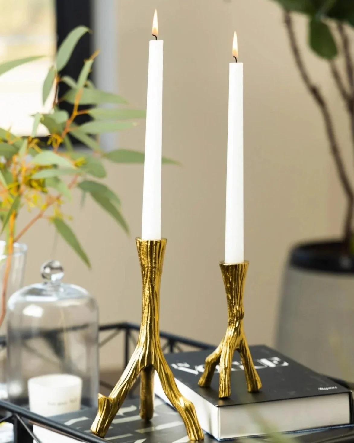 Two lit white taper candles with gold candle holders on a black tray, surrounded by plants and decorative items.