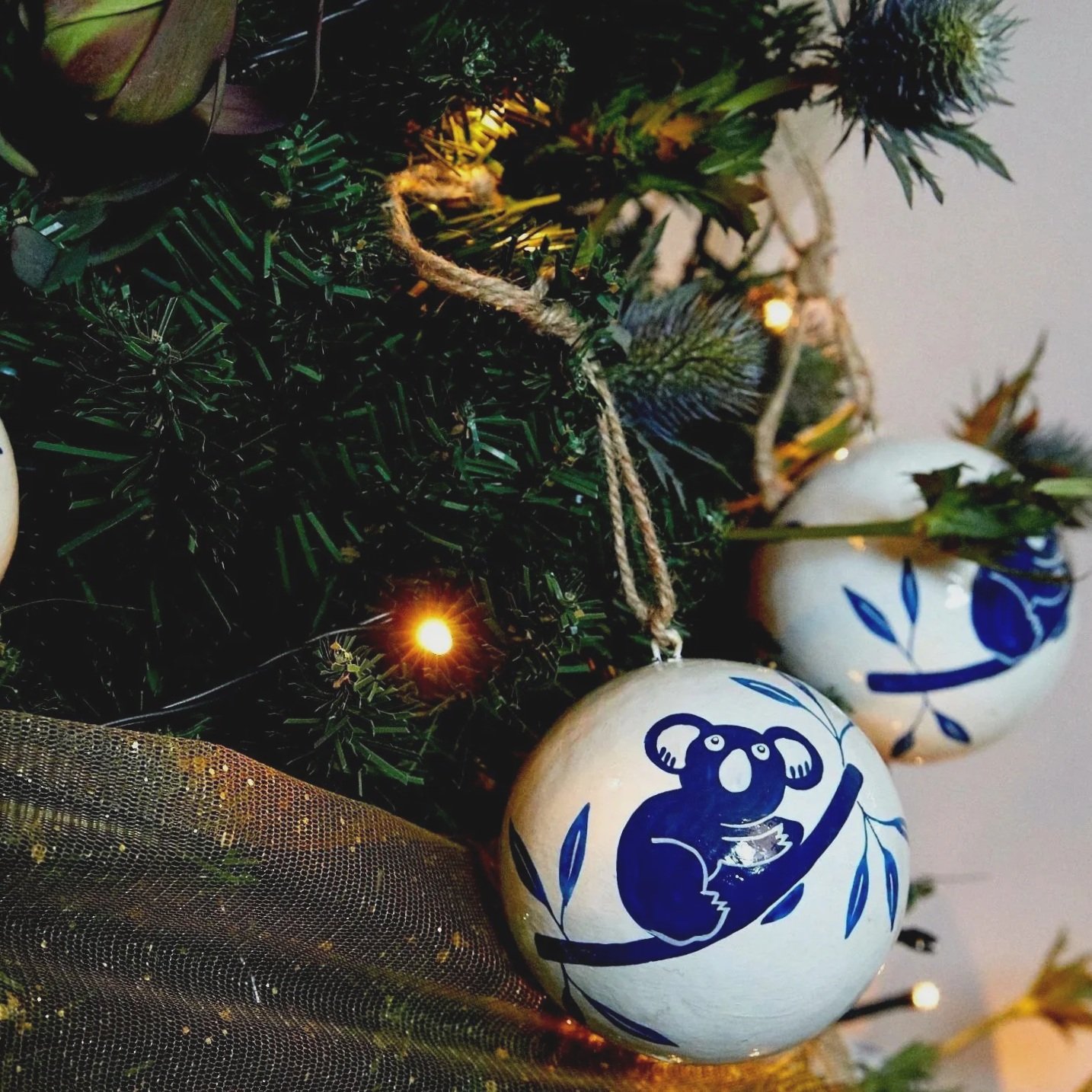 Close-up of a decorated Christmas tree with blue and white ornaments featuring a koala design, small yellow lights, and green foliage.