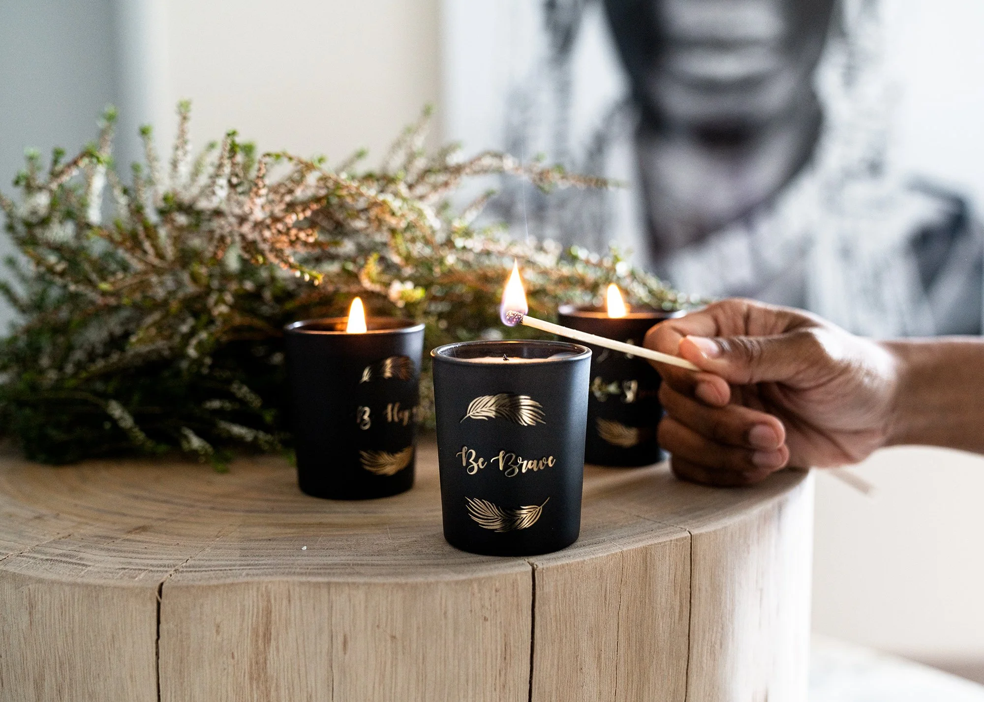 A person lighting candles in small black jars with gold feather designs and the words "Be Brave" on a wooden table, with a decorative plant in the background.