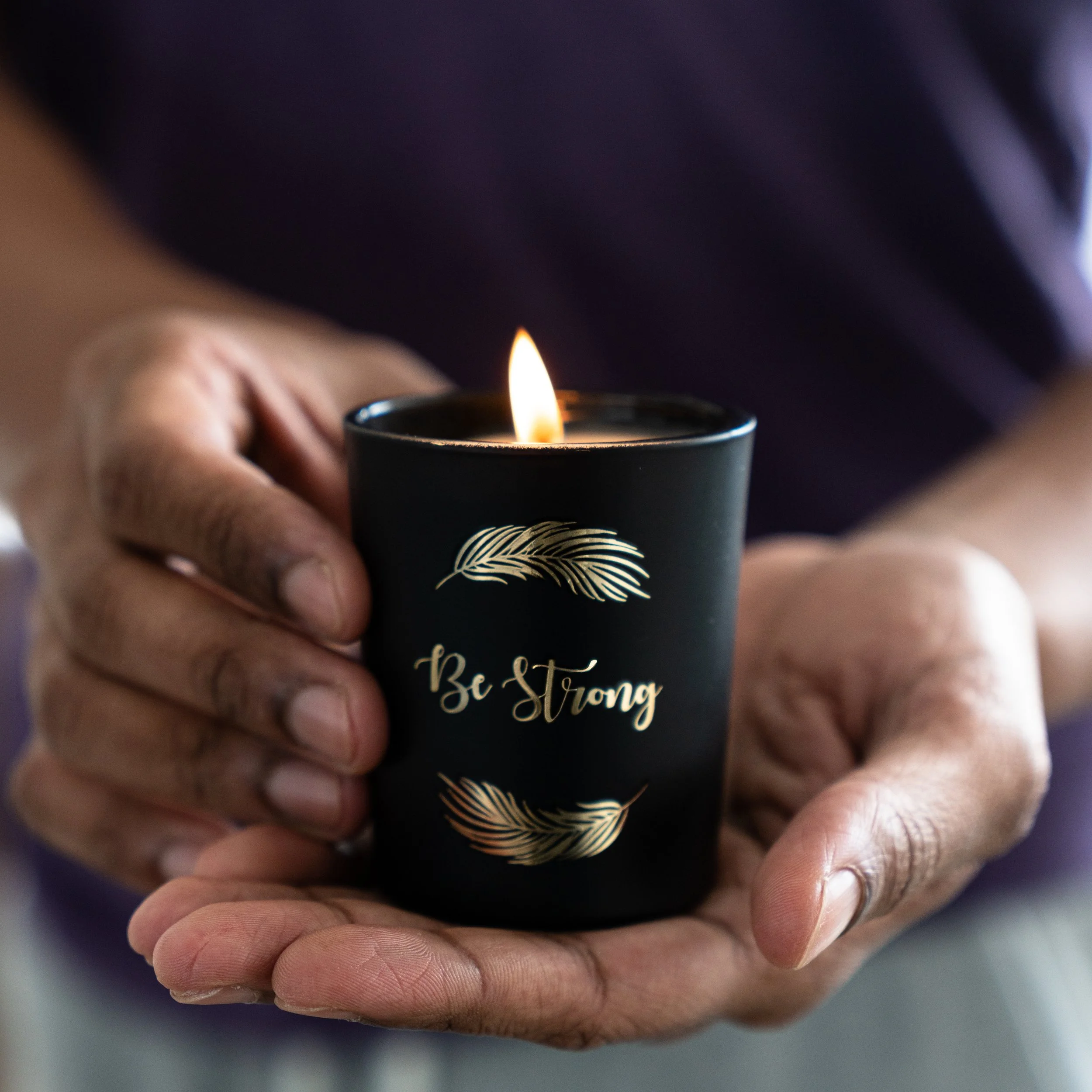 Close-up of a person's hand holding a black candle with a gold feather design and the words 'Be Strong' written on it, with a small flame burning on top.