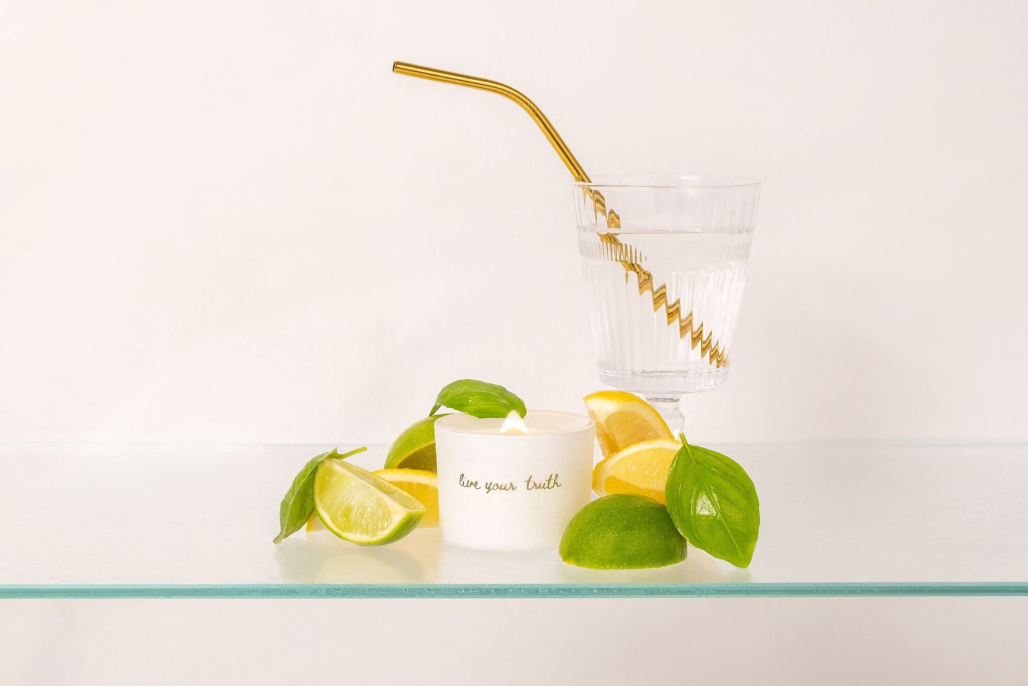 A clear glass of water with a gold metal straw, surrounded by lime wedges and green leaves, with a white candle labeled "live your truth" in the foreground.
