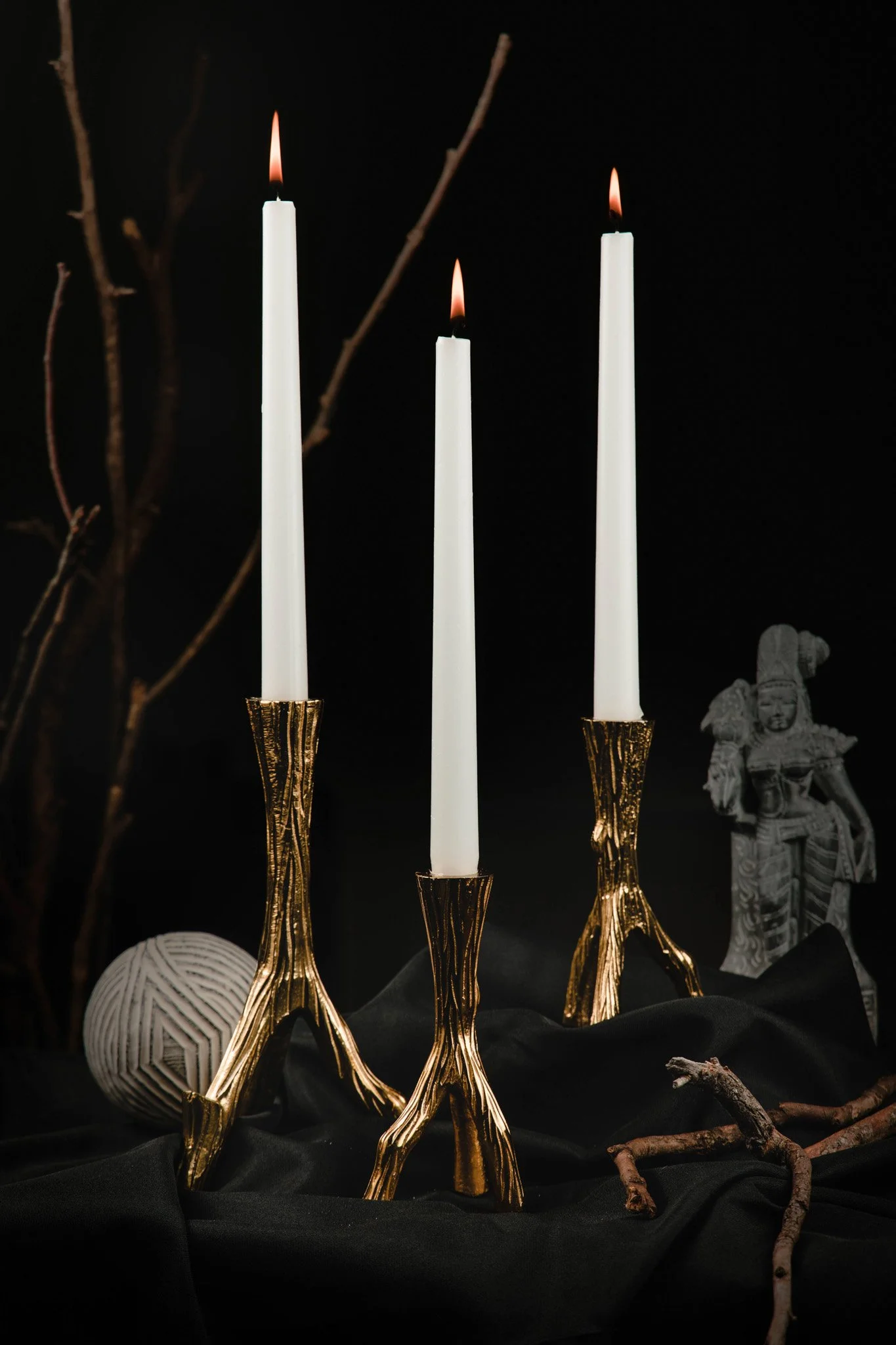 Three tall white candles with flames in gold holders, set on a black cloth, with decorative branches, a white ball, a stone sculpture of a goddess, and twigs around.