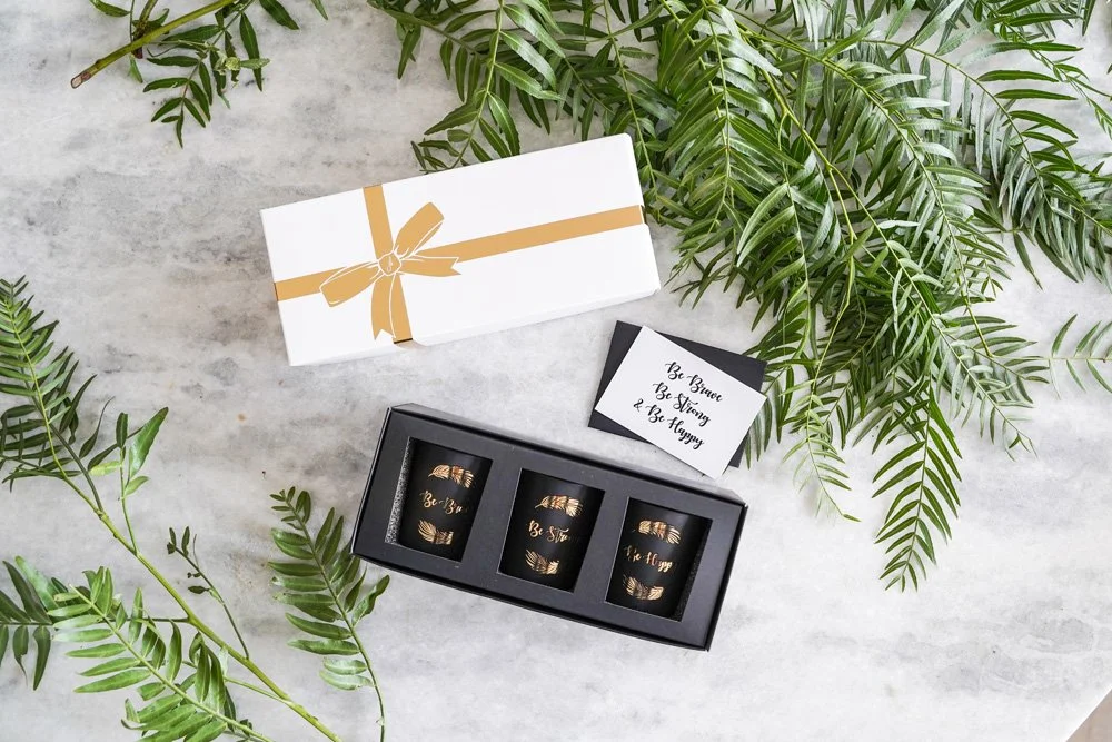 A gift box with a gold ribbon design, three glass candles with gold writing, and a motivational quote card, arranged on a marble surface with green fern leaves.