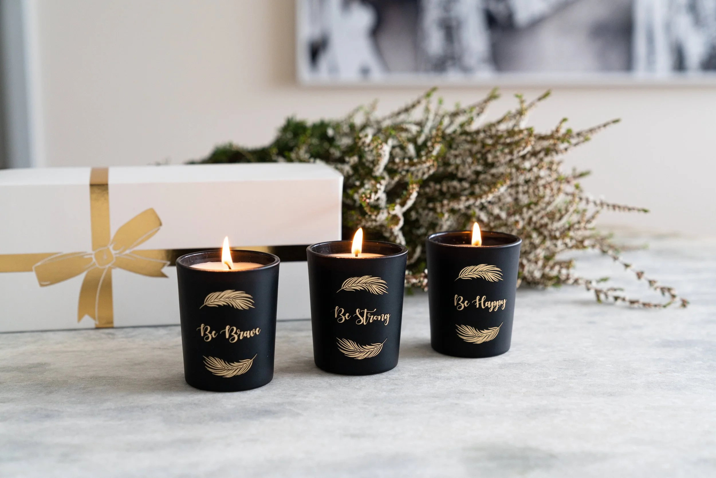 Three lit black candles with gold writing and feather designs on a white surface, with a white gift box adorned with a gold ribbon and bow, and a bouquet of white and pink flowers in the background.