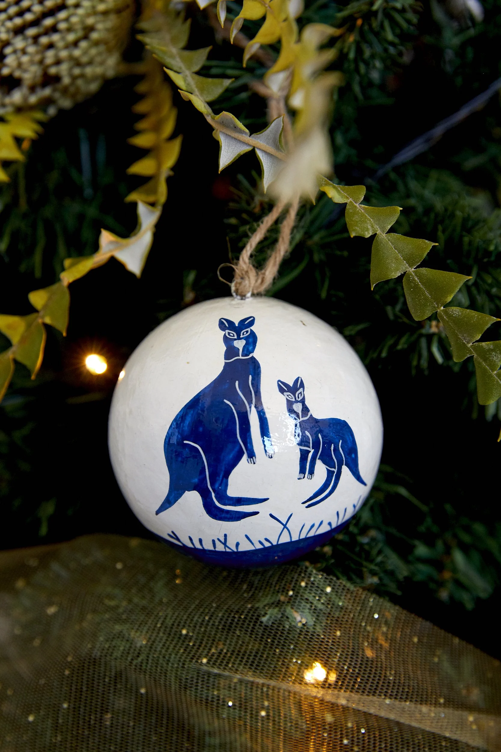 Close-up of a decorated Christmas tree with blue and white ornaments featuring a kangaroo and joey design, small yellow lights, and green foliage.


