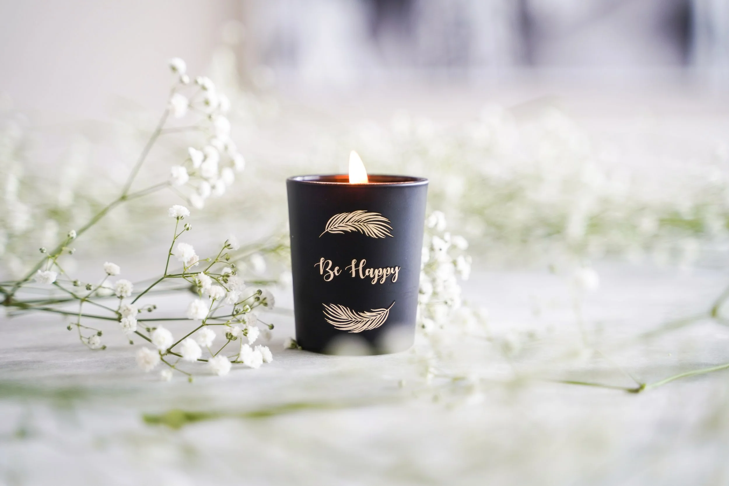 A lit black candle with a gold feather design and the words "Be Happy" written on it, surrounded by small white flowers on a light surface with a blurred background.
