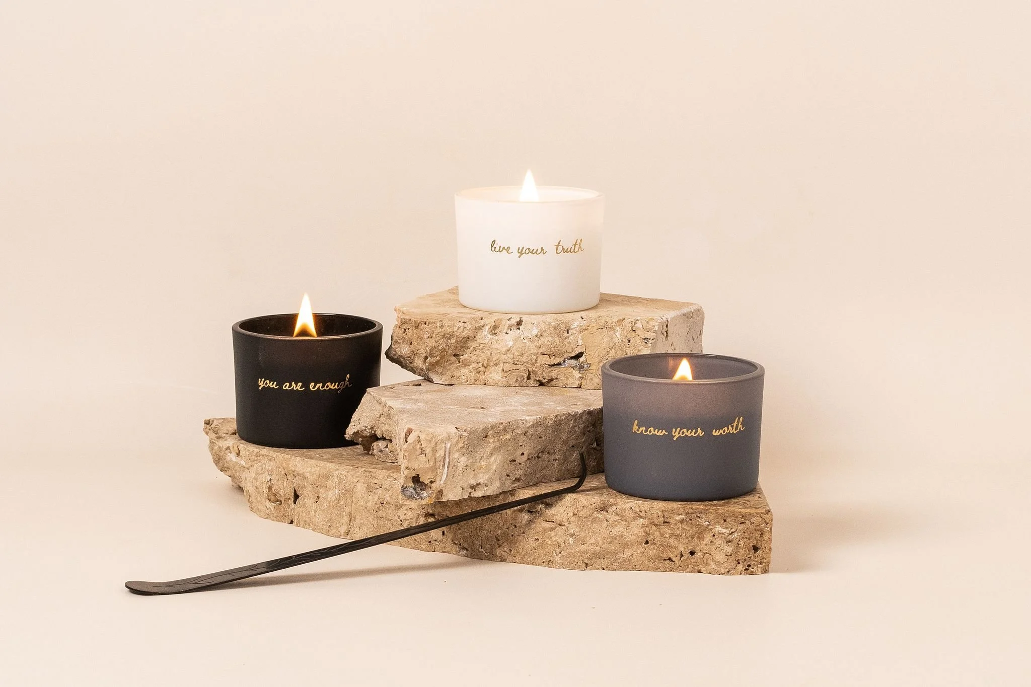 Three candles in black, white, and gray holders with gold writing, placed on a piece of rough stone, with a plain background.