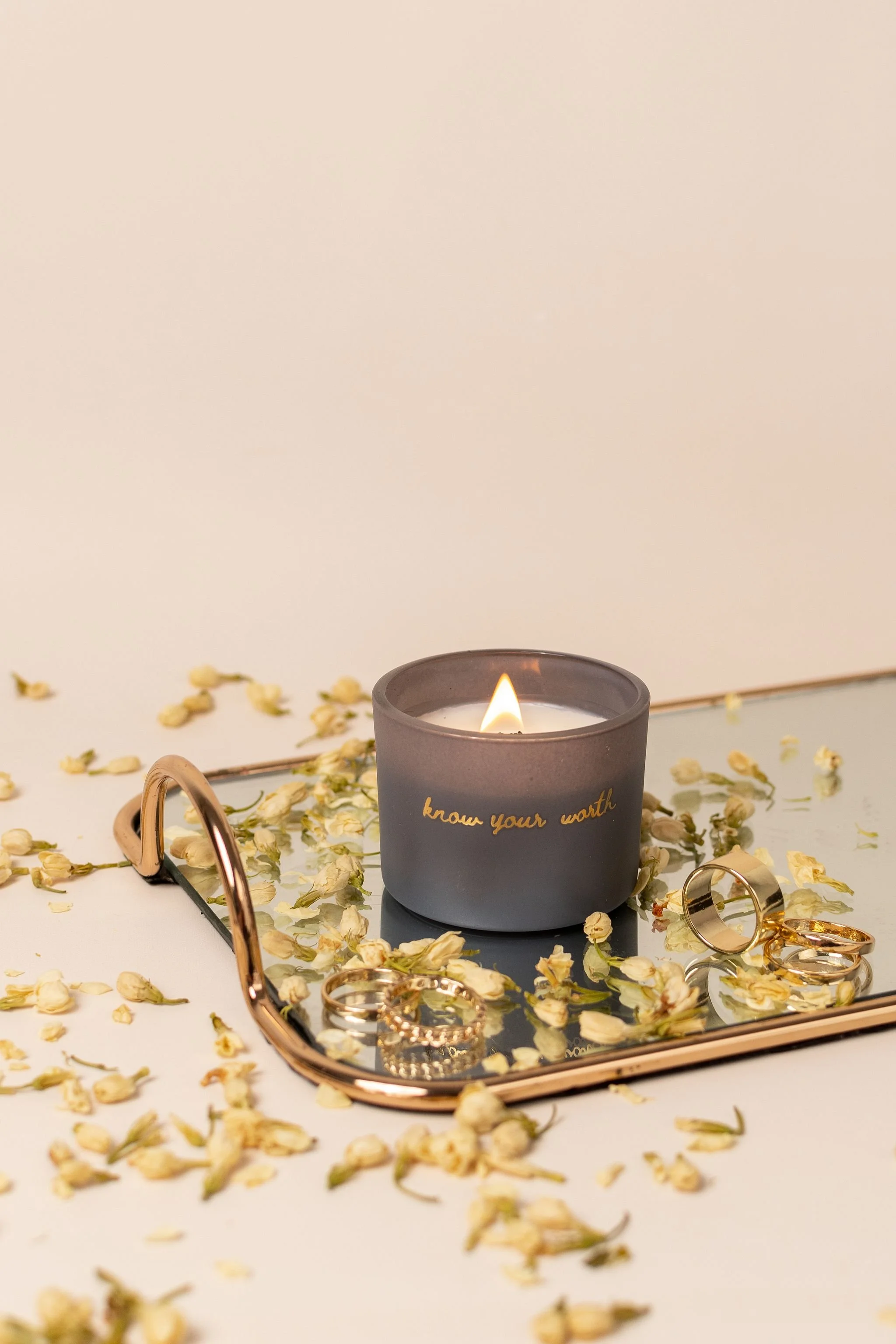 A gray candle with the text "know your worth" written on it, surrounded by scattered jasmine flowers and gold jewellery on a tray with a mirror surface