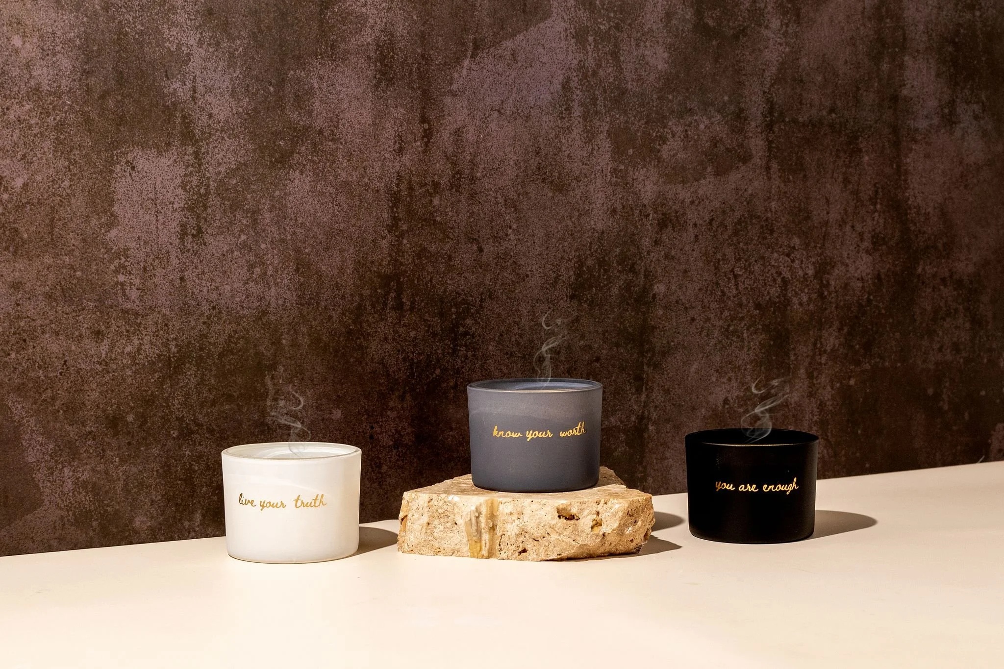 A display of three candles against a textured brown background. The candles are white, gray, and black, with gold and black writing.