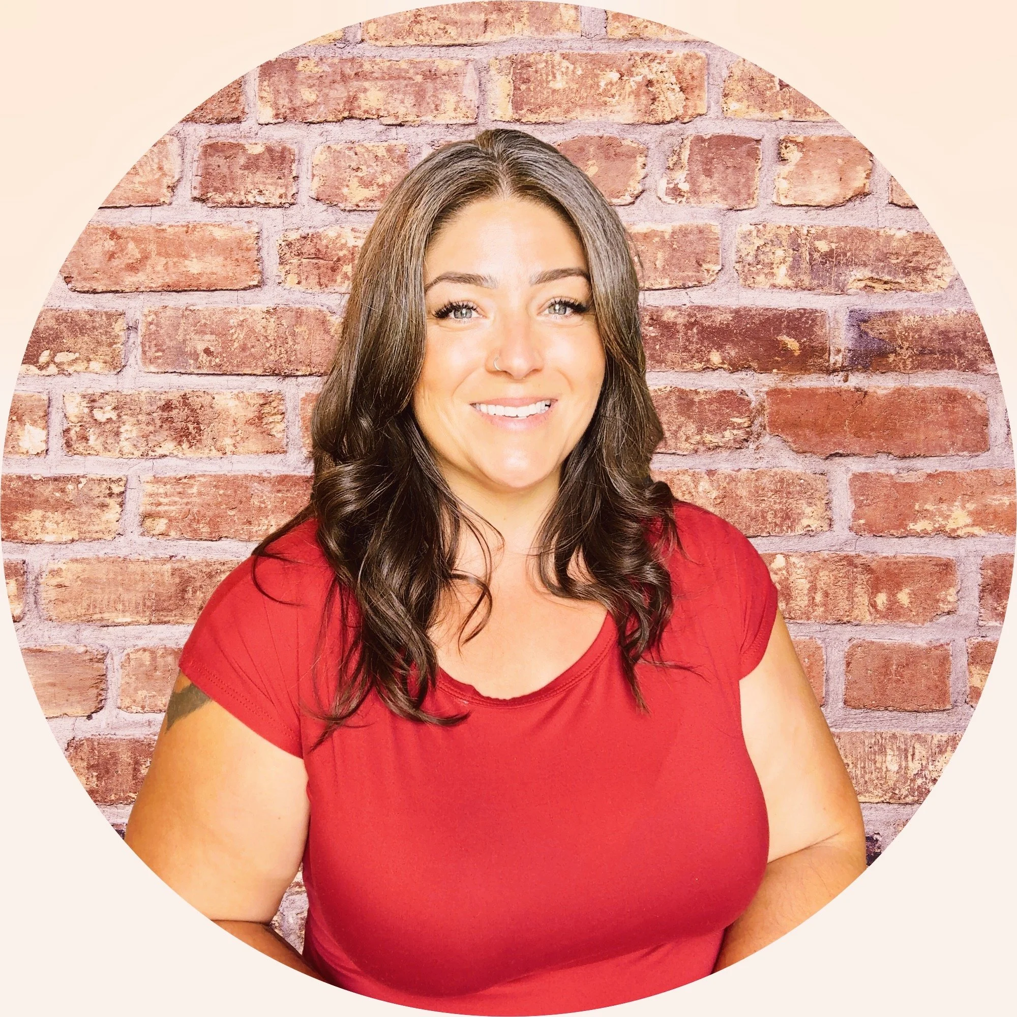 A smiling woman with long brown hair, wearing a red shirt, standing in front of a brick wall.