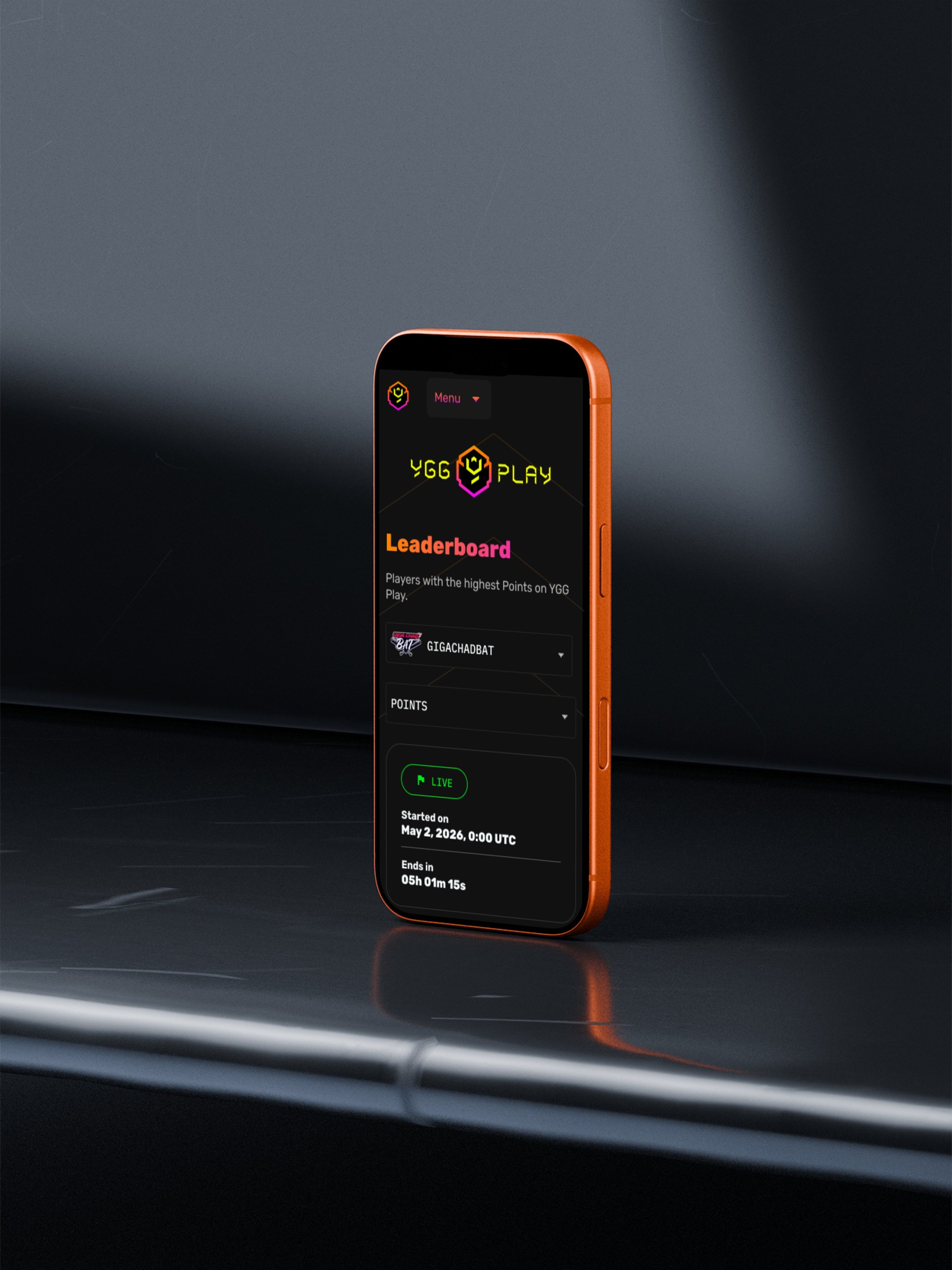 Close-up of a smartphone with an orange frame on a dark reflective surface, displaying a gaming app called YGG Play with a leaderboard, game start date, and timer, against a dark background with textured lighting.
