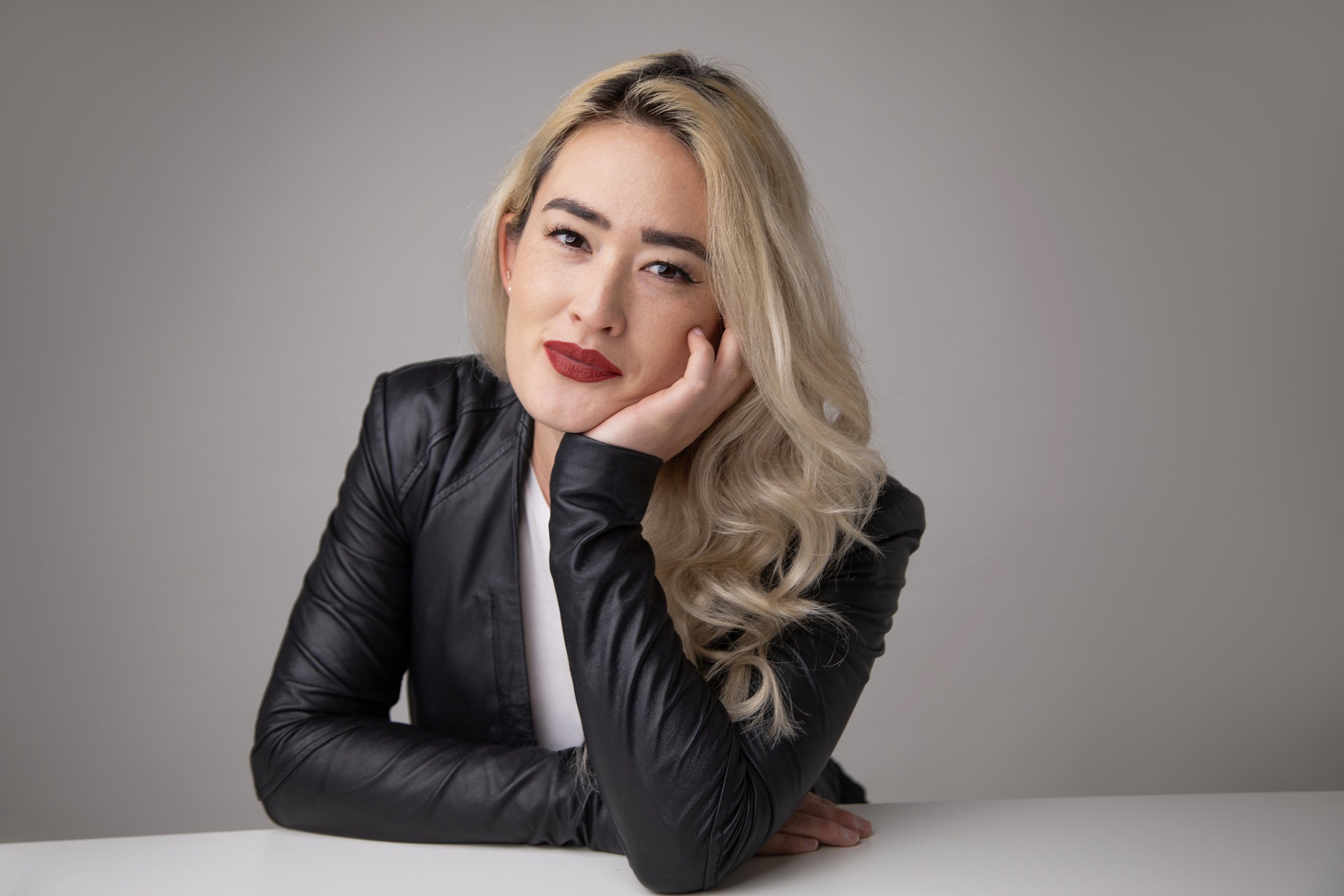 A woman with blonde wavy hair and red lipstick resting her chin on her hand, wearing a black leather jacket, sitting at a white table against a gray background.