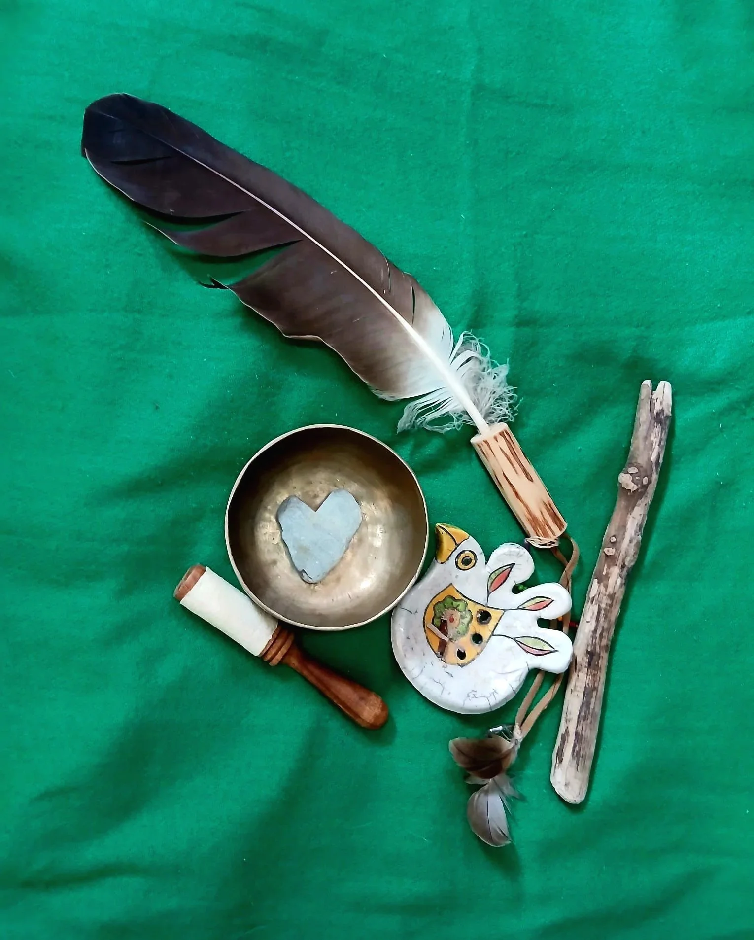 A collection of decorative items on a green fabric background, including a large black feather, a small metallic bowl with a heart shape inside, a carved wooden figure of a rabbit with painted details, and two pieces of driftwood.
