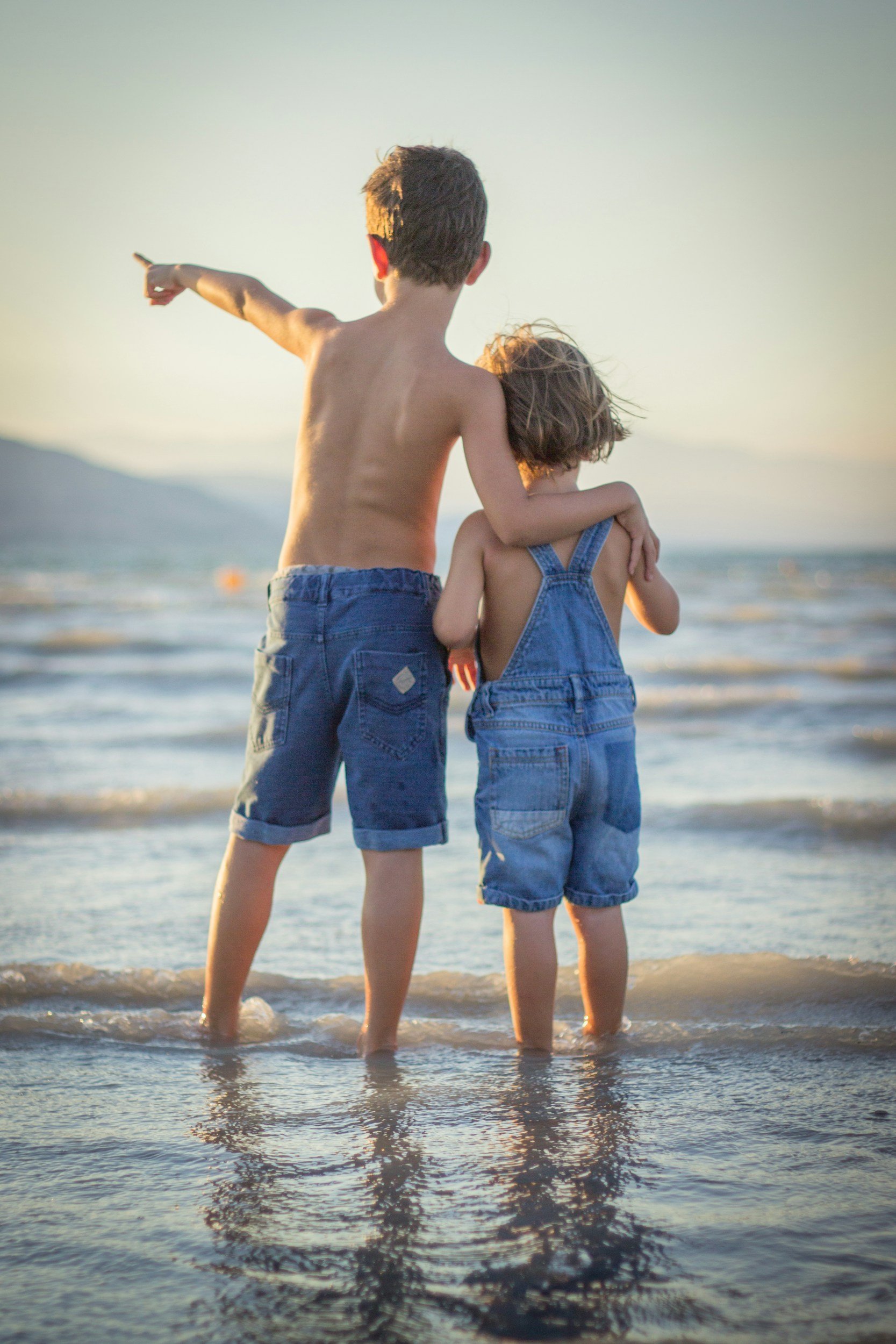 Deux enfants au bord de la mer, vu de derrière, avec la mer et le soleil couchant en arrière-plan. L'un pointe vers l'horizon, l'autre a les bras autour de l'autre.