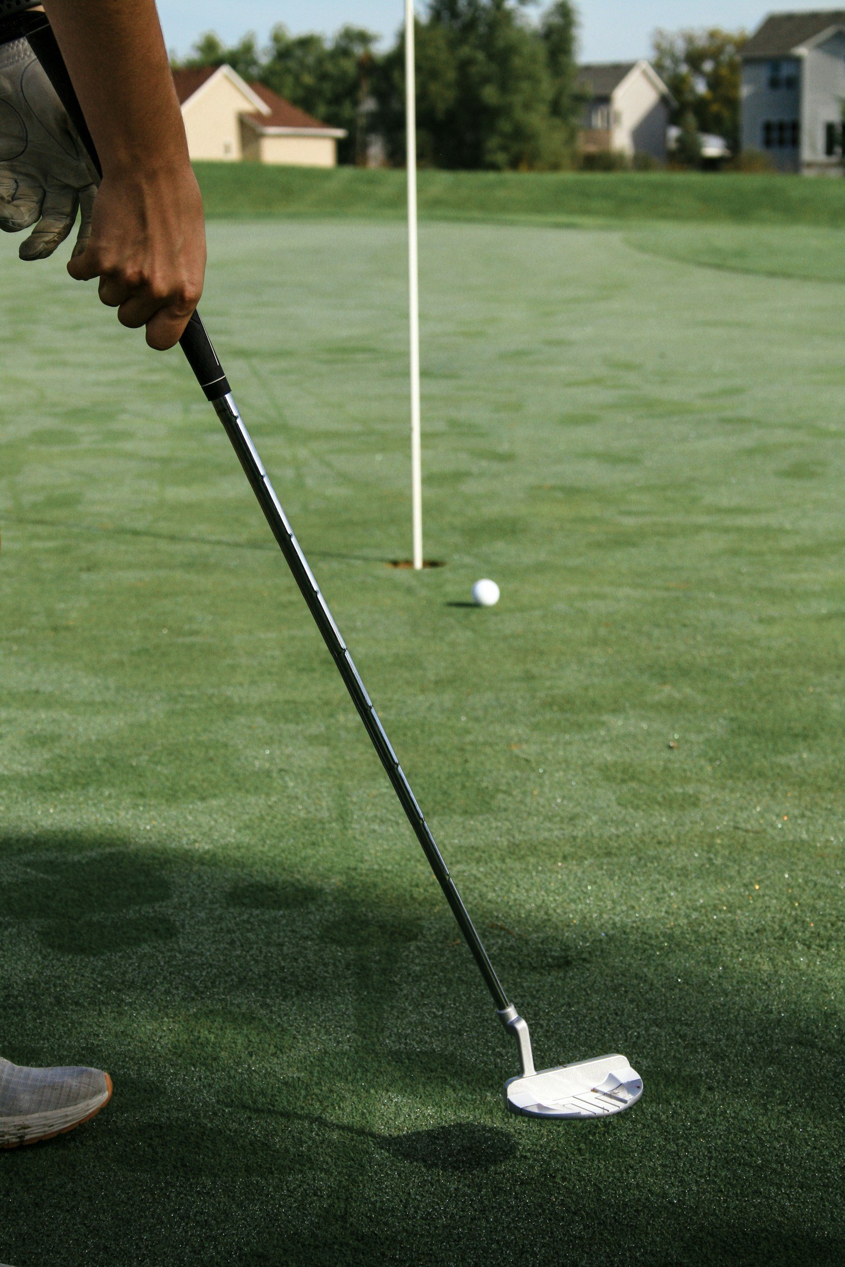 Une personne se prépare à taper un coup de golf sur un parcours de golf vert avec des maisons en arrière-plan, un drapeau blanc marque l'emplacement du trou.